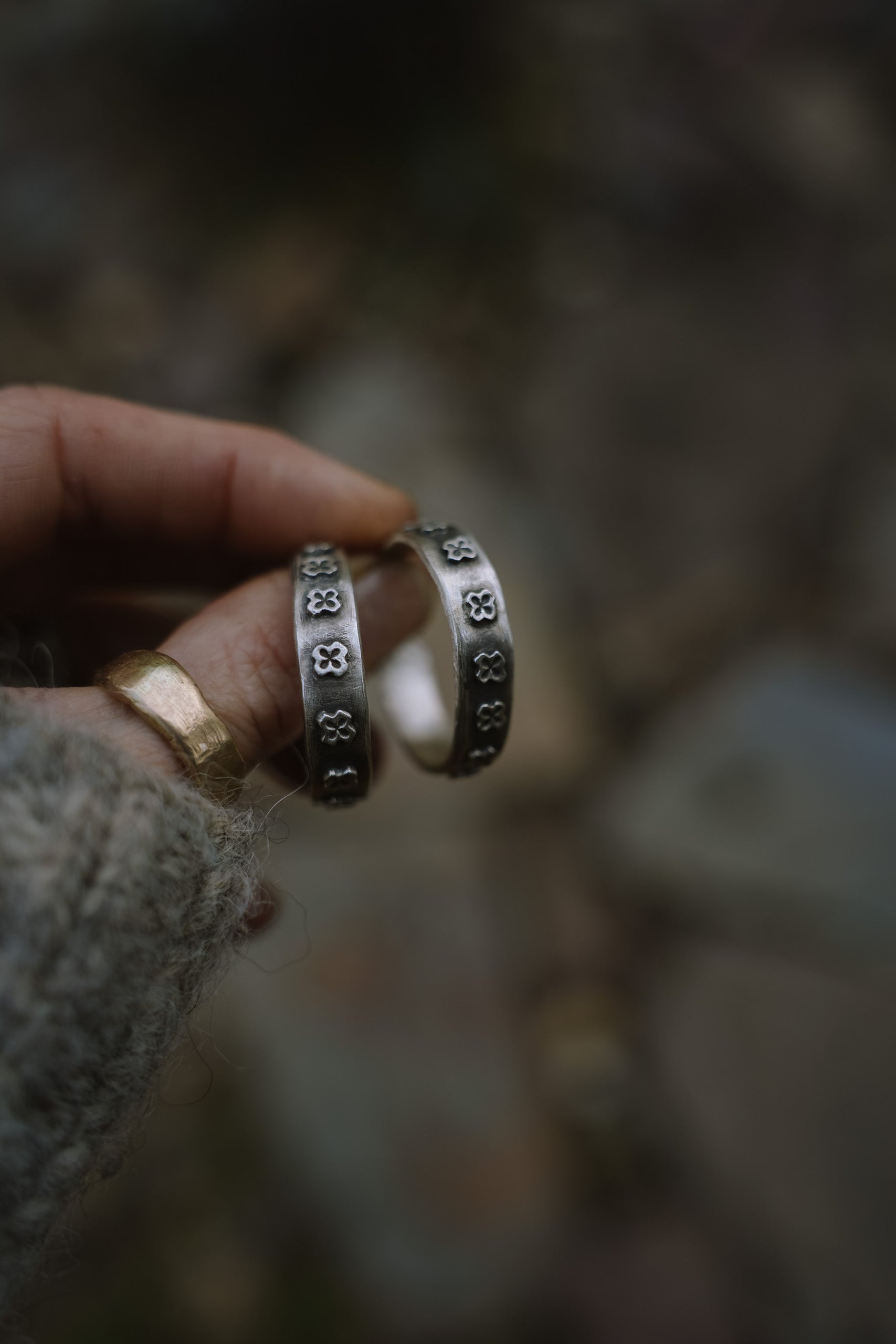 Close-up of a woman's hand holding a pair of large, wide-band sterling silver hoop earrings. The earrings are adorned with raised, hand-carved small crosses. The silver is oxidized, creating, in some areas, a contrast of black against silver. The natural background is somewhat dark and blurred.