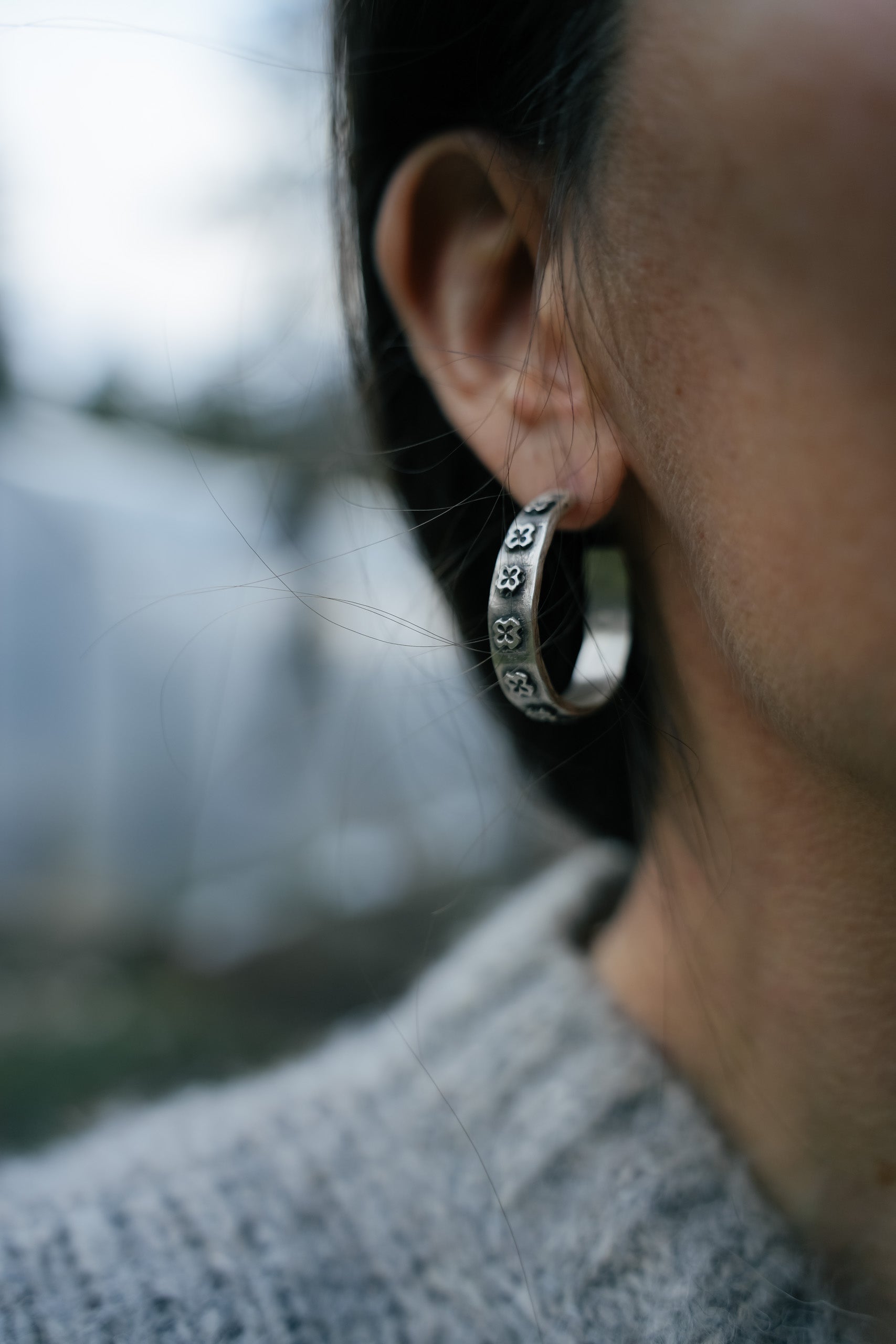 Close-up of a woman's ear with a large, wide-band sterling silver hoop earring. The earring body is adorned with raised, hand-carved small crosses. The silver is oxidized, creating, in some areas, a contrast of black against silver. She is wearing a gray sweater and has long black hair.