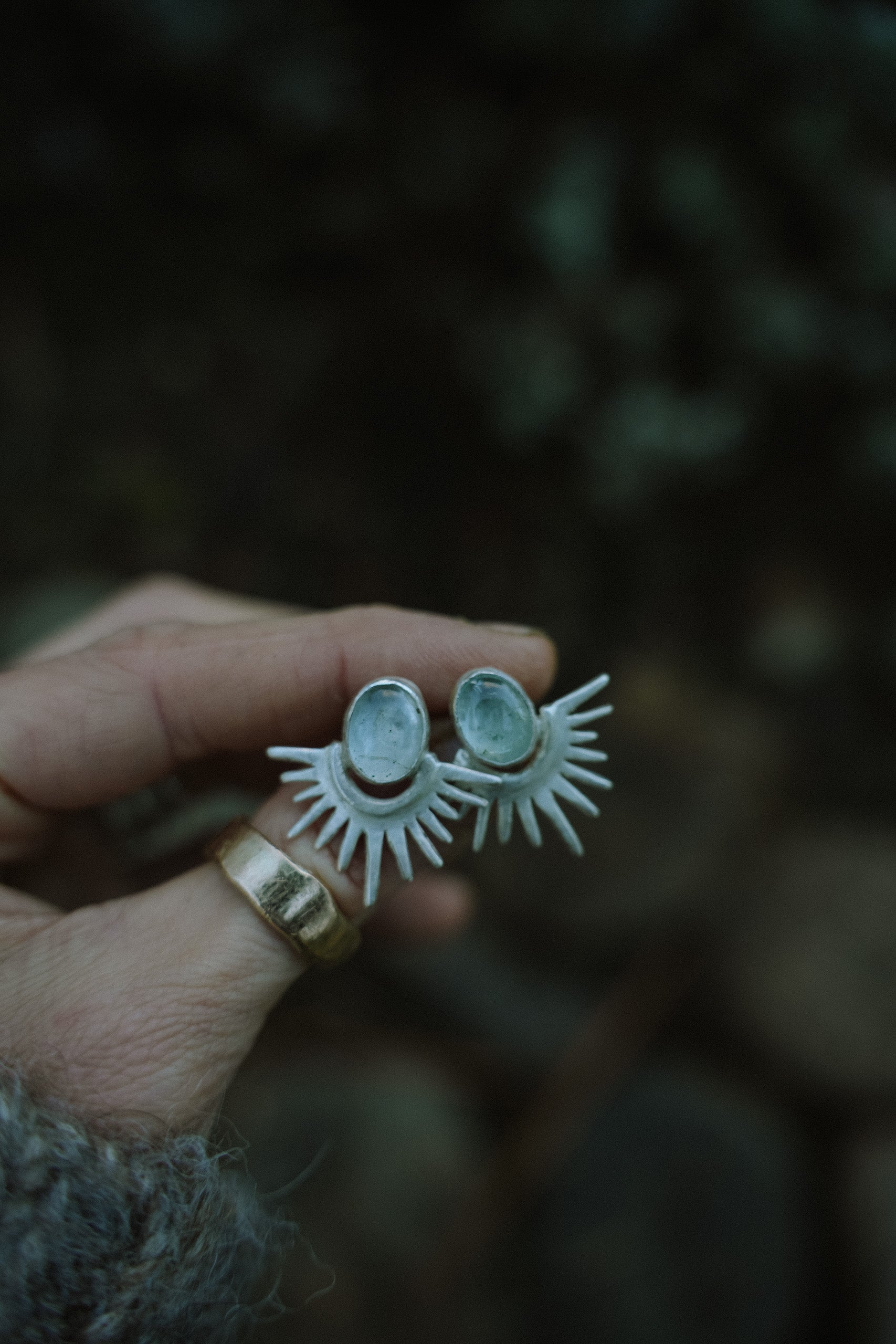 A woman's hand hand holding a pair of sterling silver and aquamarine post earrings agains a dark background. The silver rays resemble the reflection of the sun’s rays on a lake with the turquoise orbs illustrating the sun.