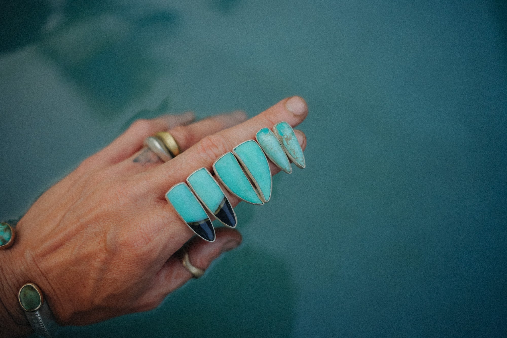 A woman's hand holding three pairs of sterling silver and chrysocolla talon-shaped, post earrings against a teal background