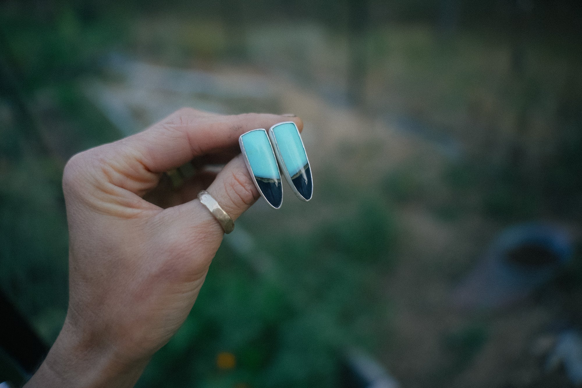 A woman's hand holding a pair of  two-toned chrysocolla gemstones and sterling silver, talon-shaped, post-style earrings against a blurred natural background
