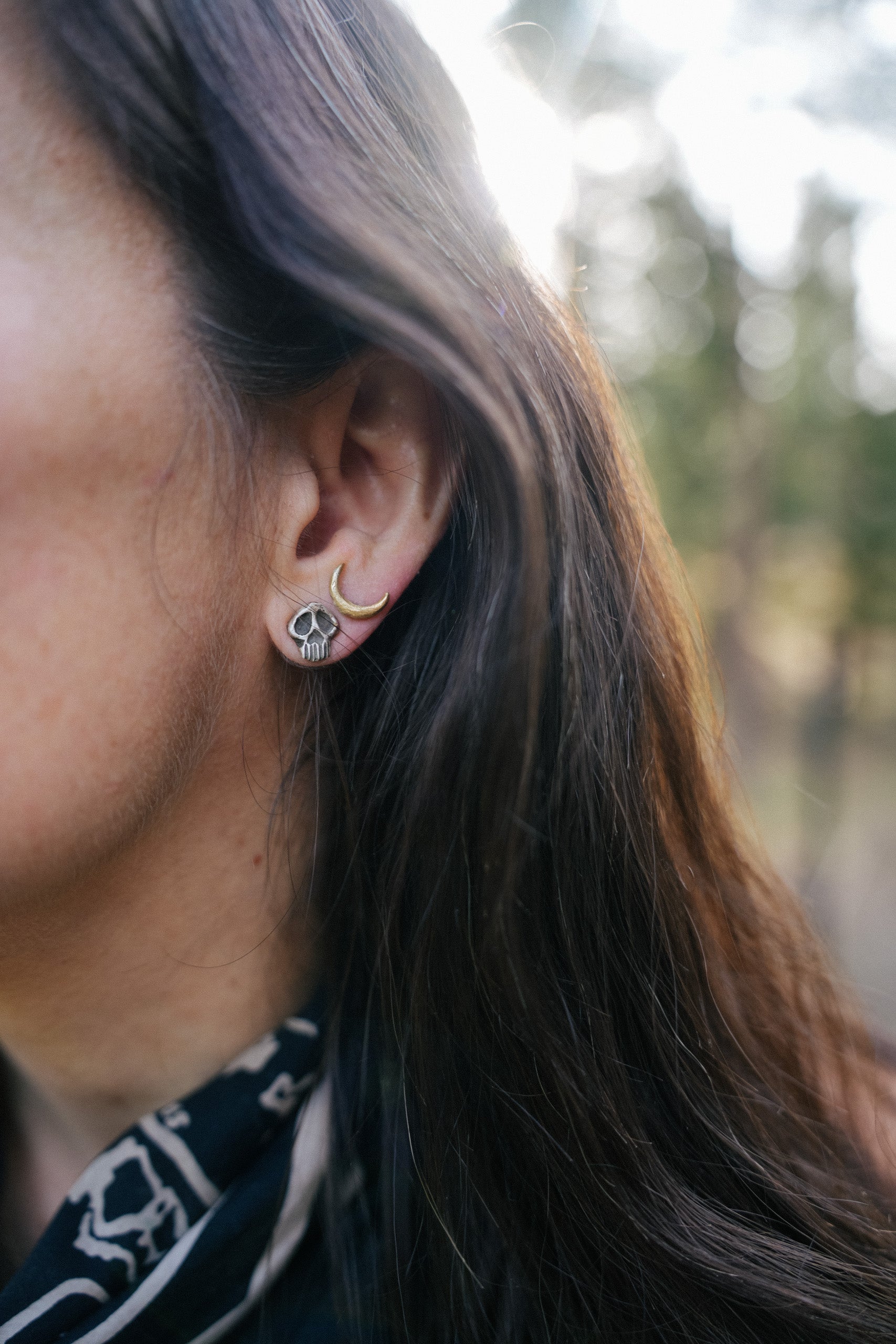 A woman wearing a small sterling silver Momento Mori post earring, along with a slim 18K gold crescent-shaped post earring. She has long, dark hair.