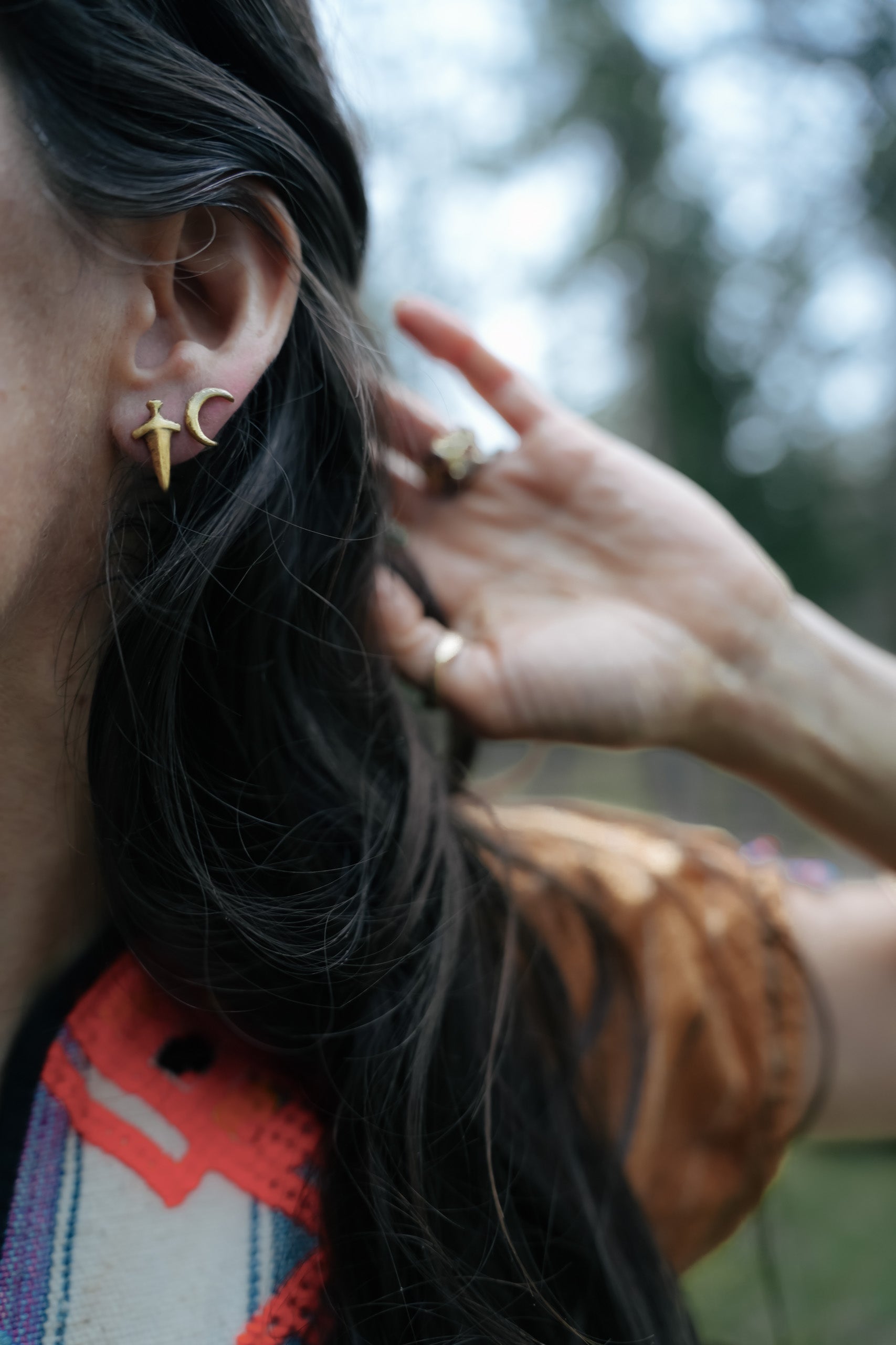 Close-up of a woman with long dark hair wearing a 14K gold post earring shaped like a crescent moon. She is also wearing a 14K gold post earring shaped like a dagger.