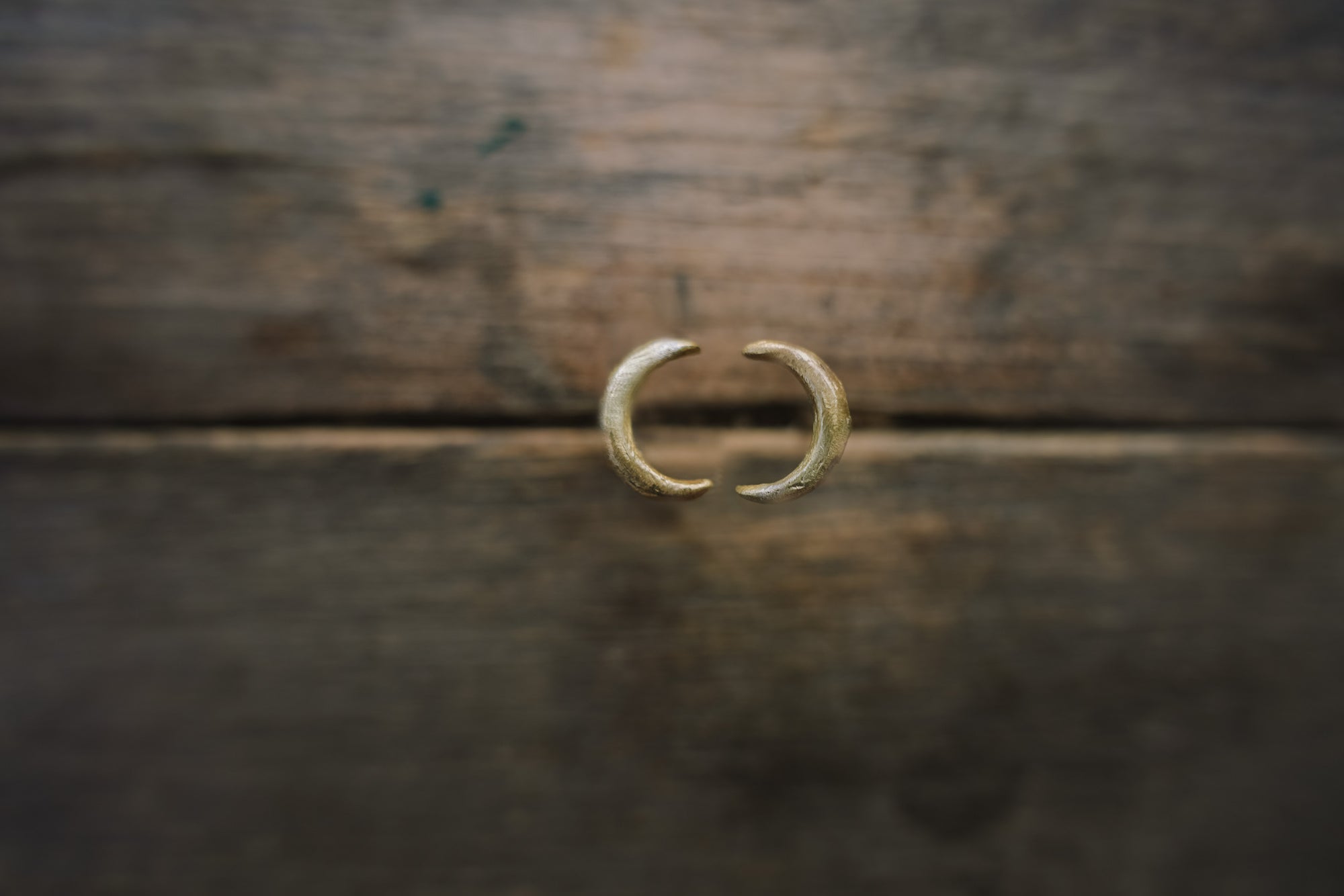 Close-up photograph of two hand-forged and carved 14K gold post earrings. They are shaped like crescent moons and have a slightly rough texture showing the carving marks.