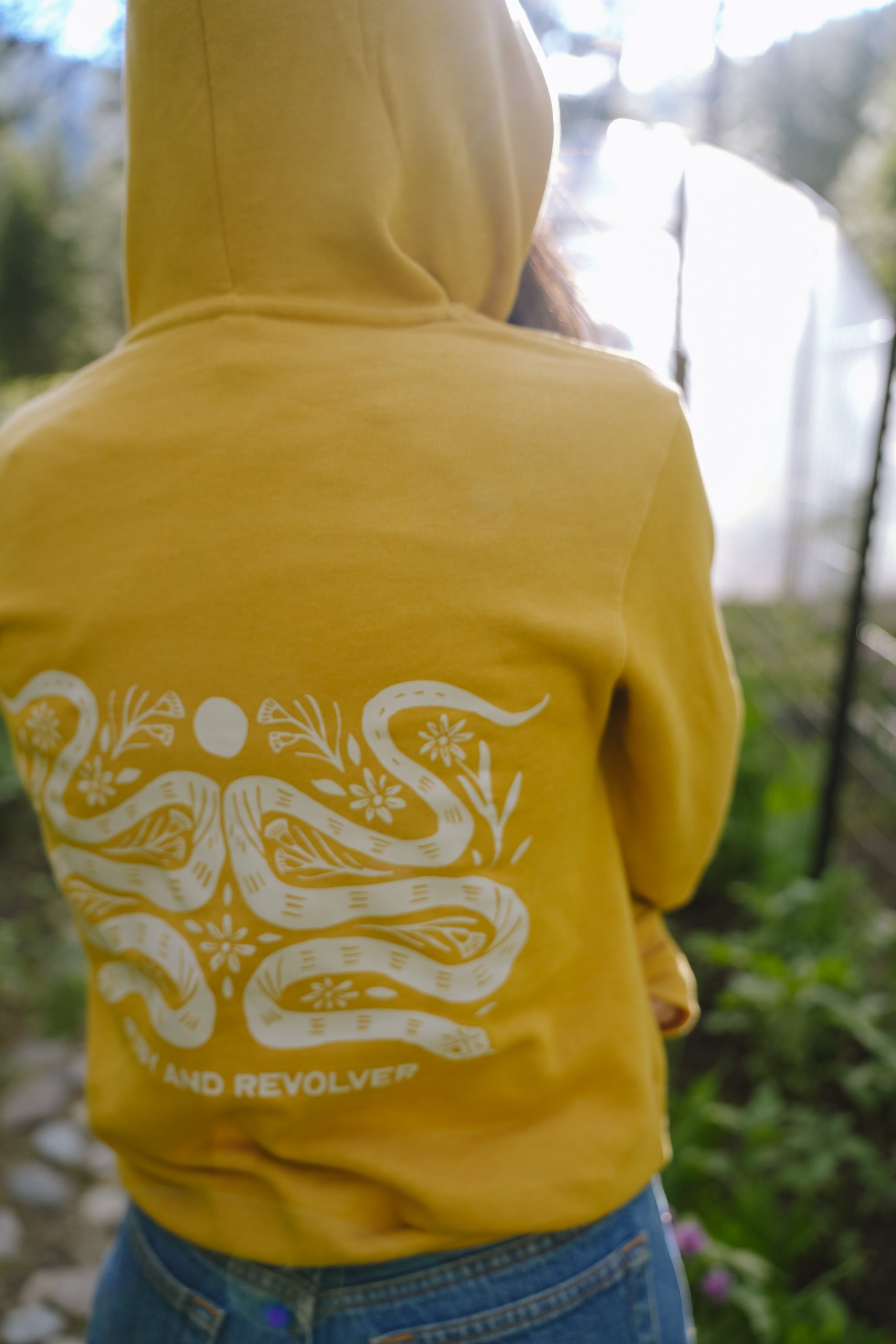 A woman, viewed from behind, wearing a mustard-yellow hoodie with a large white graphic featuring two snakes, a moon symbol, and flower icons. The text reads 'RUBY AND REVOLVER' across the back, just beneath the snakes. She is wearing blue jeans and is standing in an outdoor setting.