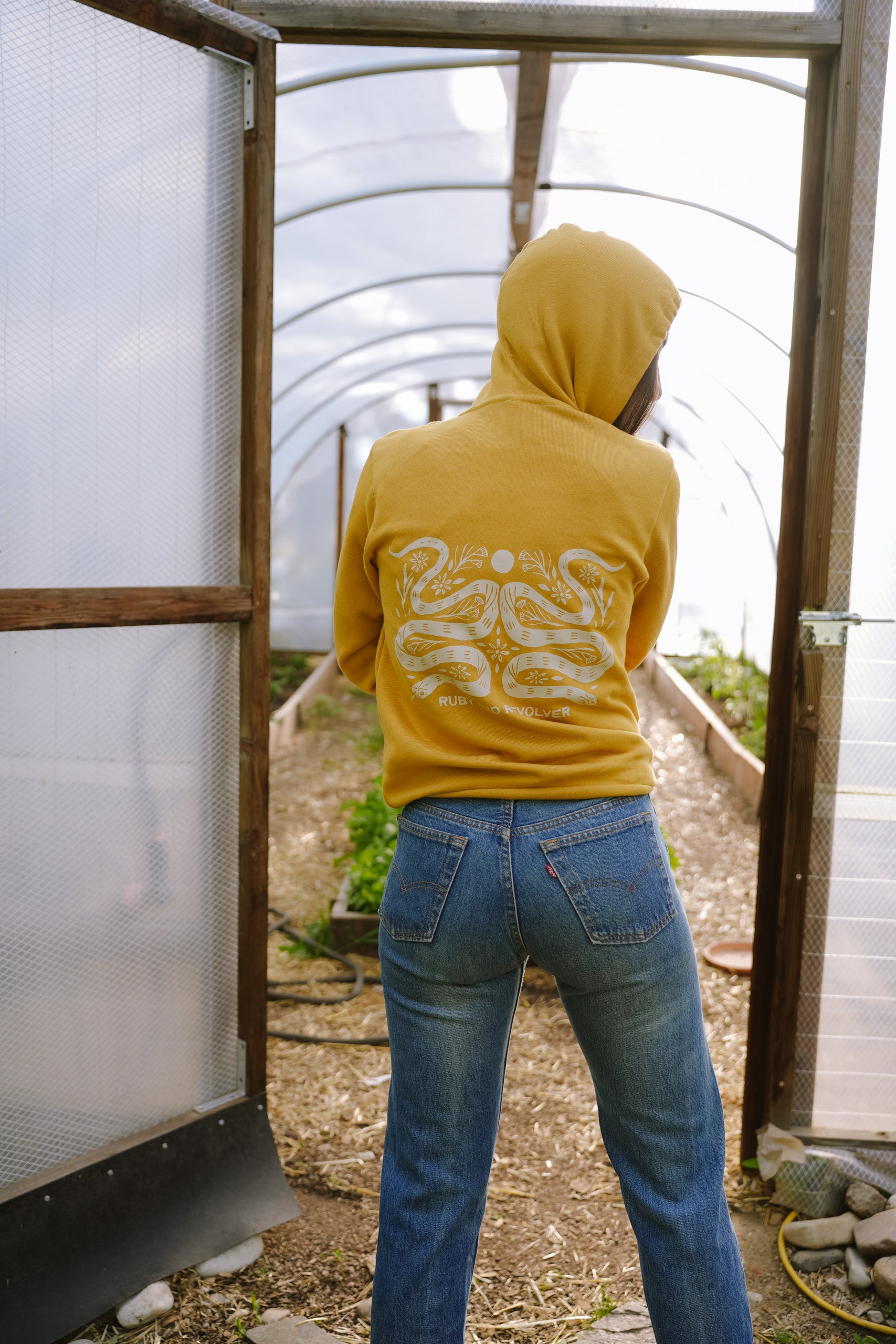 A woman, viewed from behind, wearing a mustard-yellow hoodie with a large white graphic featuring two snakes, a moon symbol, and flower icons. The text reads 'RUBY AND REVOLVER' across the back, just beneath the snakes. She is wearing blue jeans and is standing art the entrance of a hoop house.