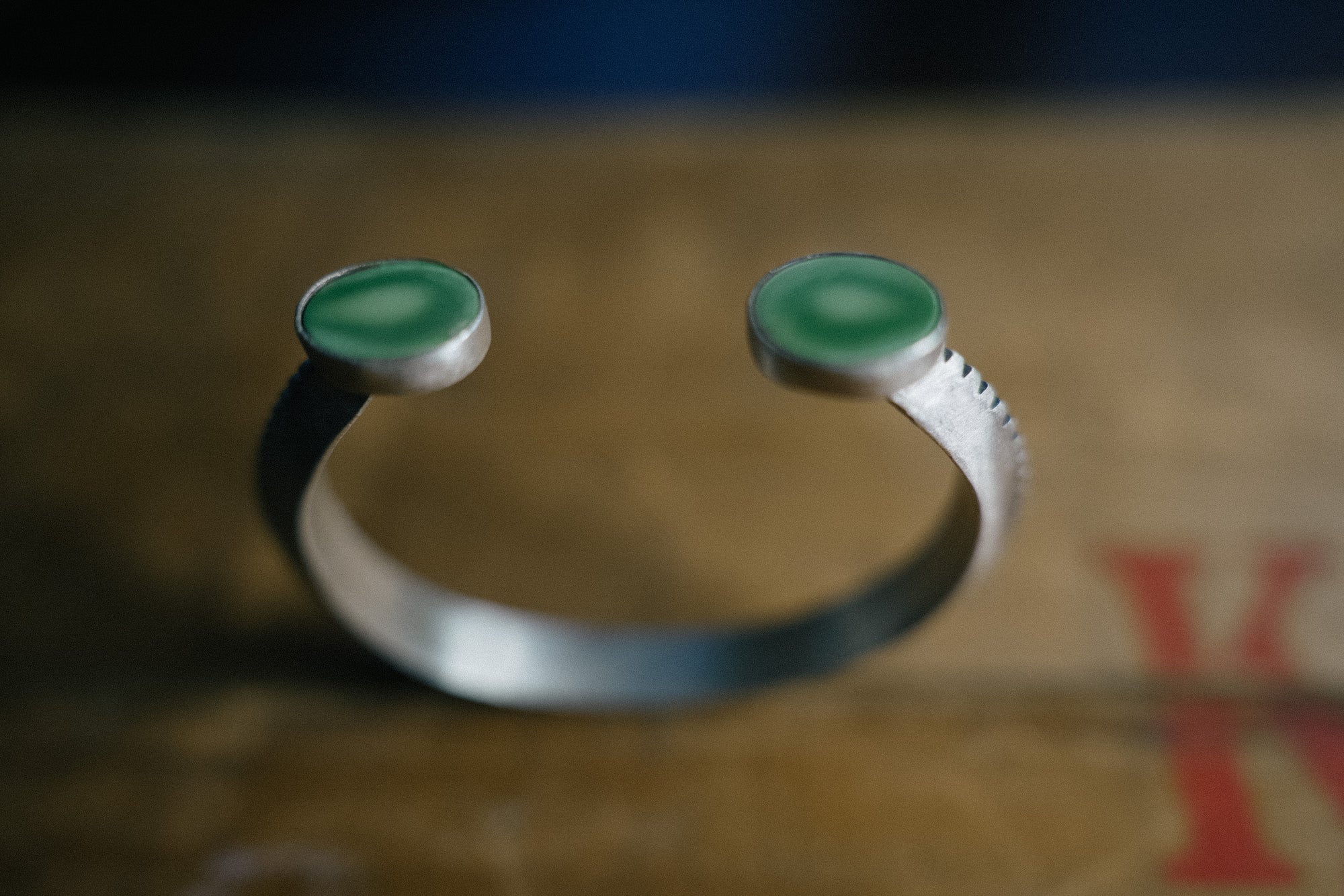 A sterling silver cuff bracelet with a serrated edge on one side and oval green turquoise gemstones at each end balances on top of a brown background.