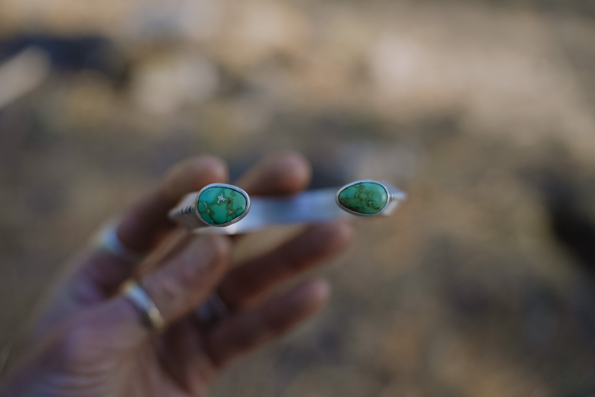 A woman's hand holds a sterling silver cuff bracelet that has tear-drop-shaped Sonoron turquoise gemstones on each end against a blurred natural background.