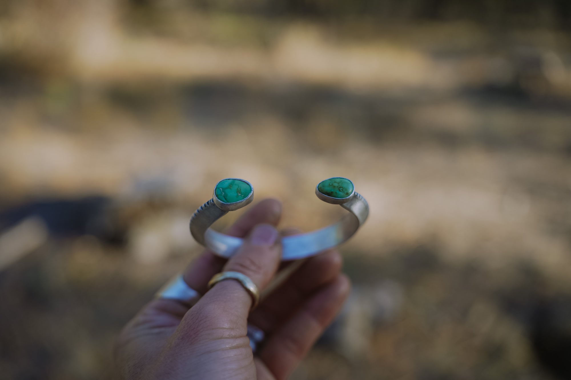 A woman's hand holds a sterling silver cuff bracelet that has tear-drop-shaped Sonoron turquoise gemstones on each end against a blurred natural background.