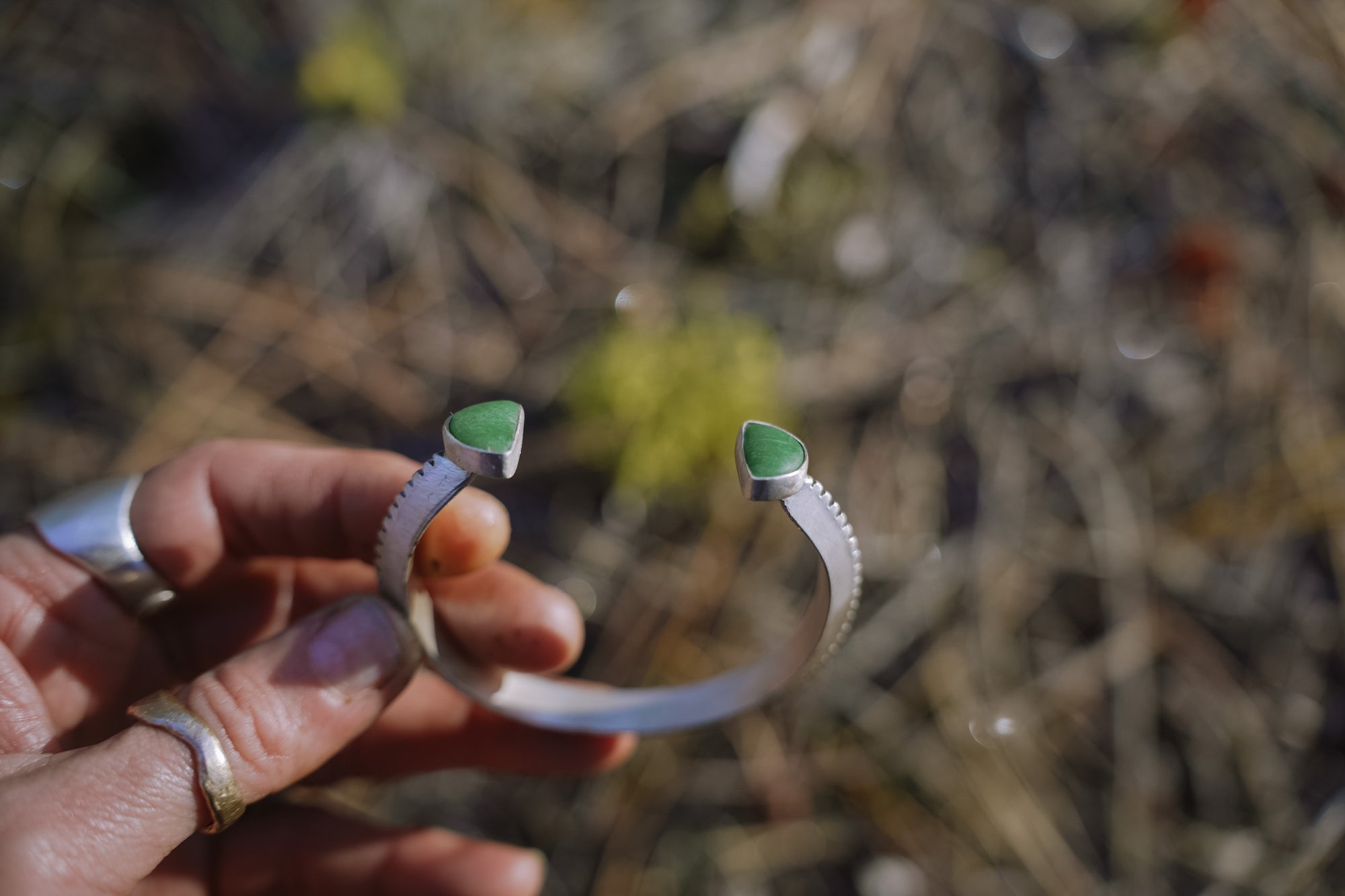 A woman's hand holding a sterling silver bracelet with green variscite gemstones at each end against a blurred natural background