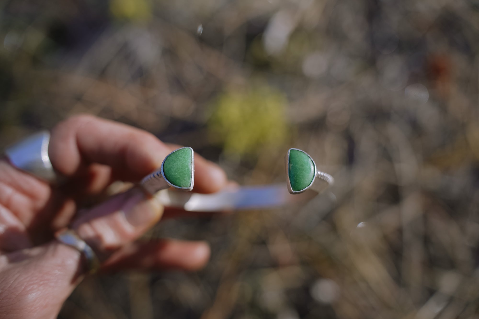 A woman's hand holding a sterling silver and green variscite gemstone bracelet against a blurred natural background. The gemstones are half-moon-shaped.