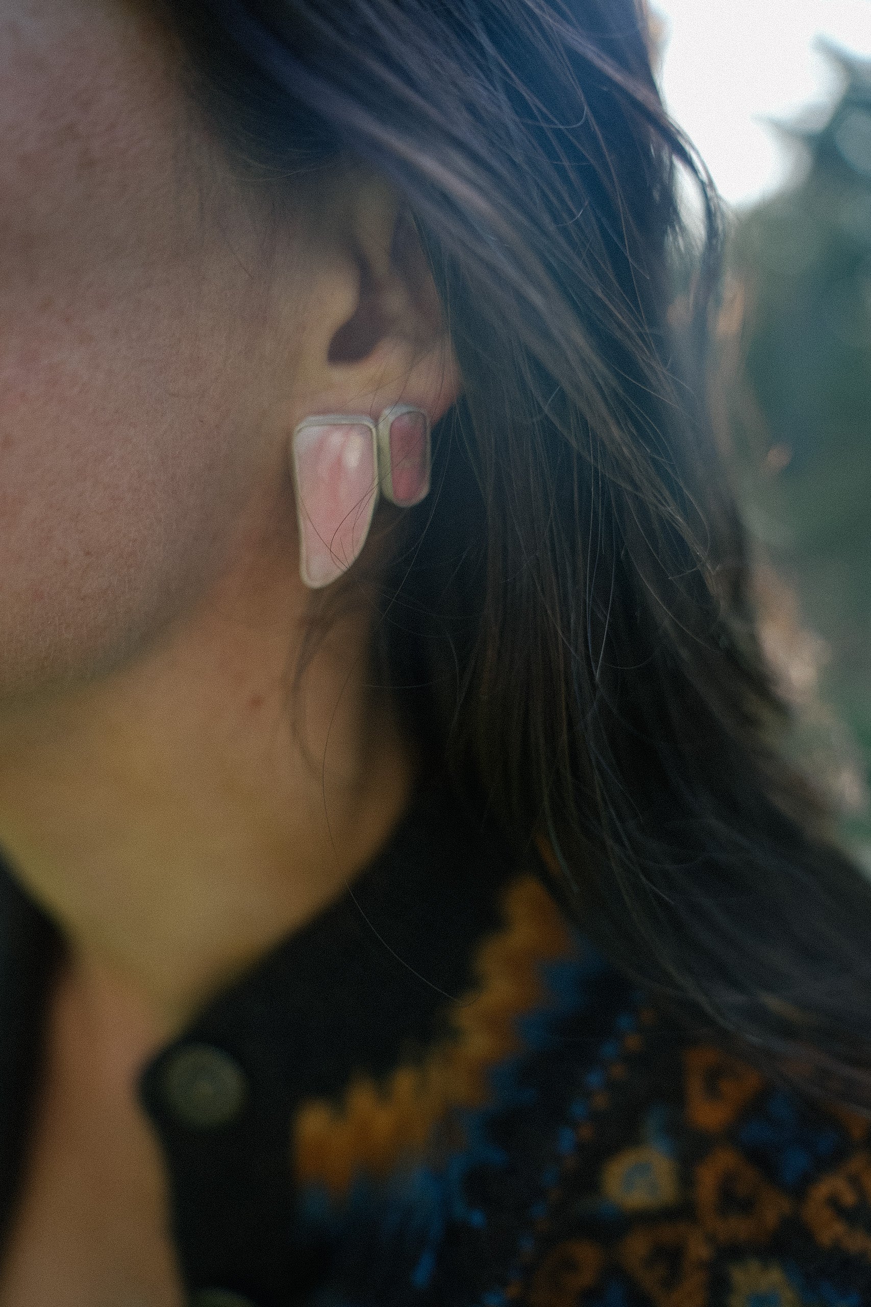 Close-up of a woman wearing pink earrings with a blurred background. The earring in front is talon-shaped and the earring behind it is bar-shaped. Both are made with sterling silver and pink opal.