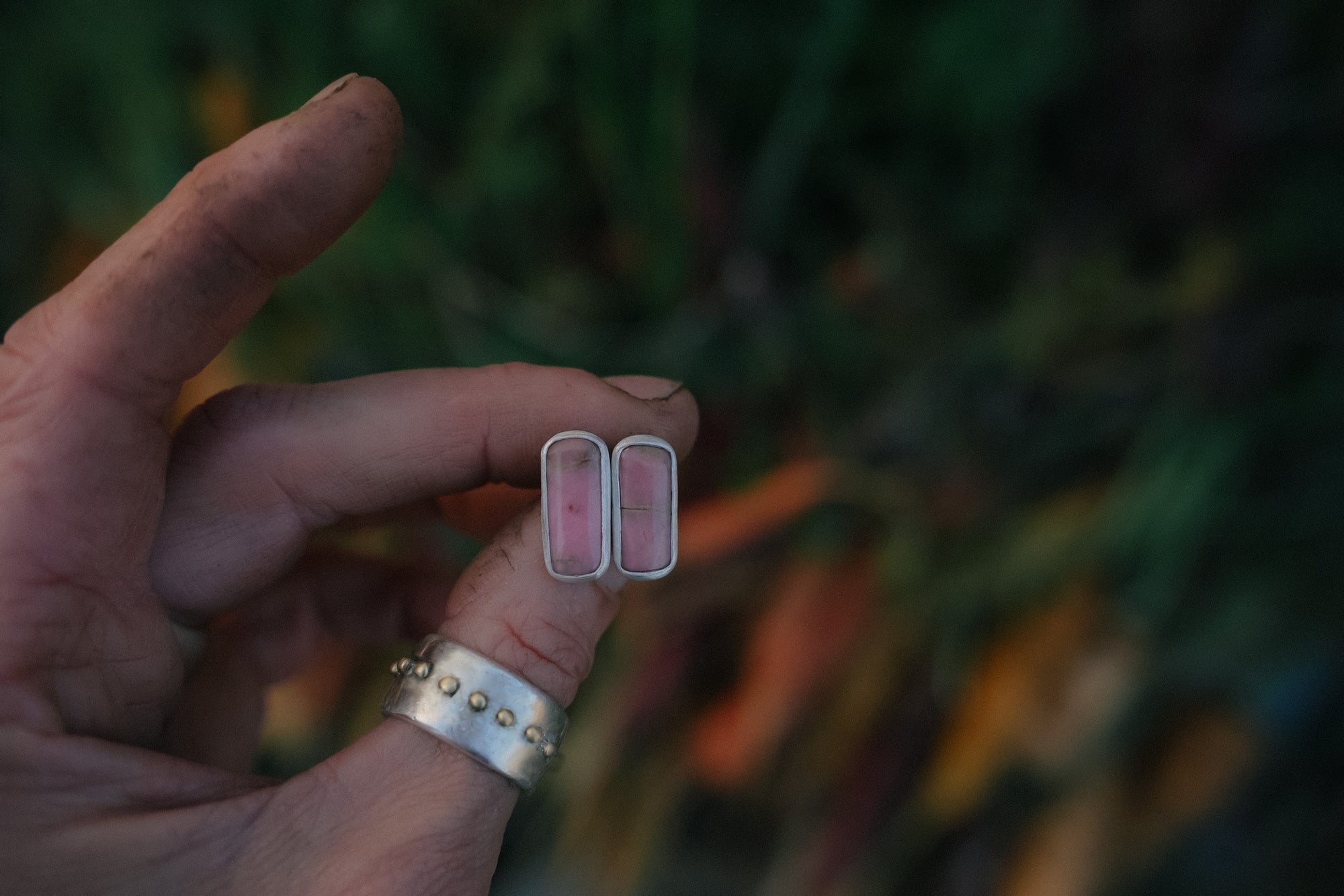 Woman;s hand holding a pair of sterling silver nad pink opal bar-shaped earrings  against a blurred background.
