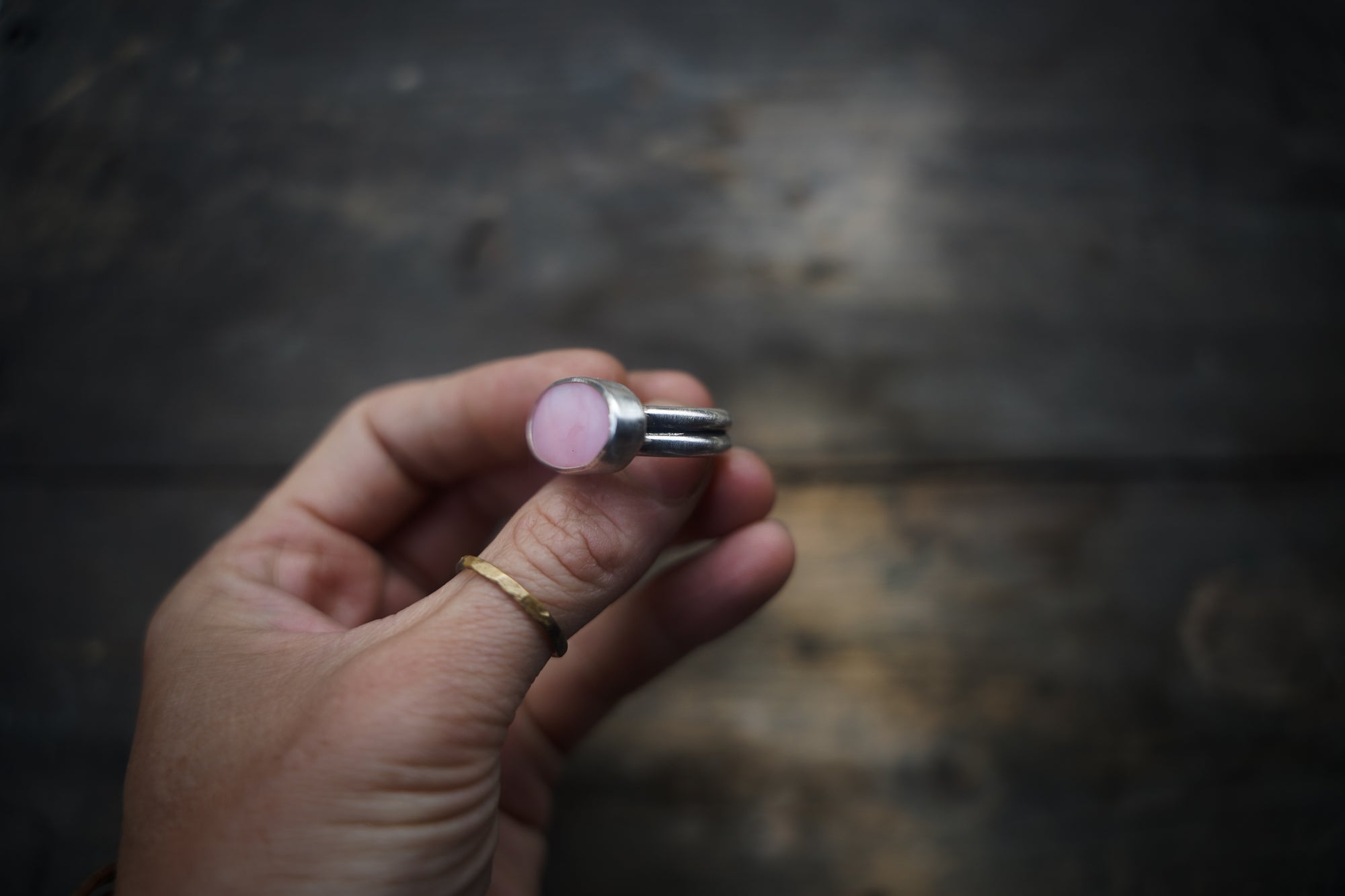 A close-up of a woman’s hand holding a pink opal and sterling silver rings, turned to show the side view. The opal is oval-shaped and set horizontally on the band. The band is made of two bands fused together to make one wider band. The natural wood background is dark and out-of-focus.