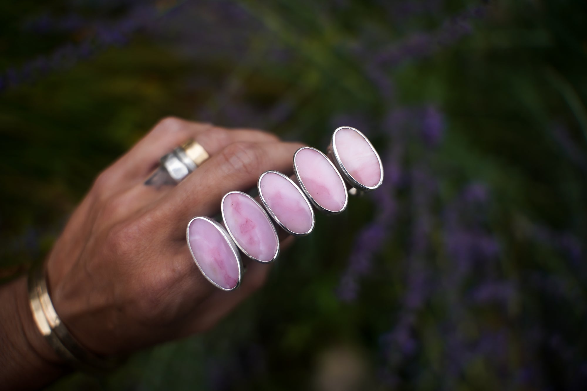A close-up of a woman’s hand wearing five pink opal and sterling silver rings on her forefinger. The opals are oval-shaped and set horizontally on the bands. The natural background is dark and out-of-focus.