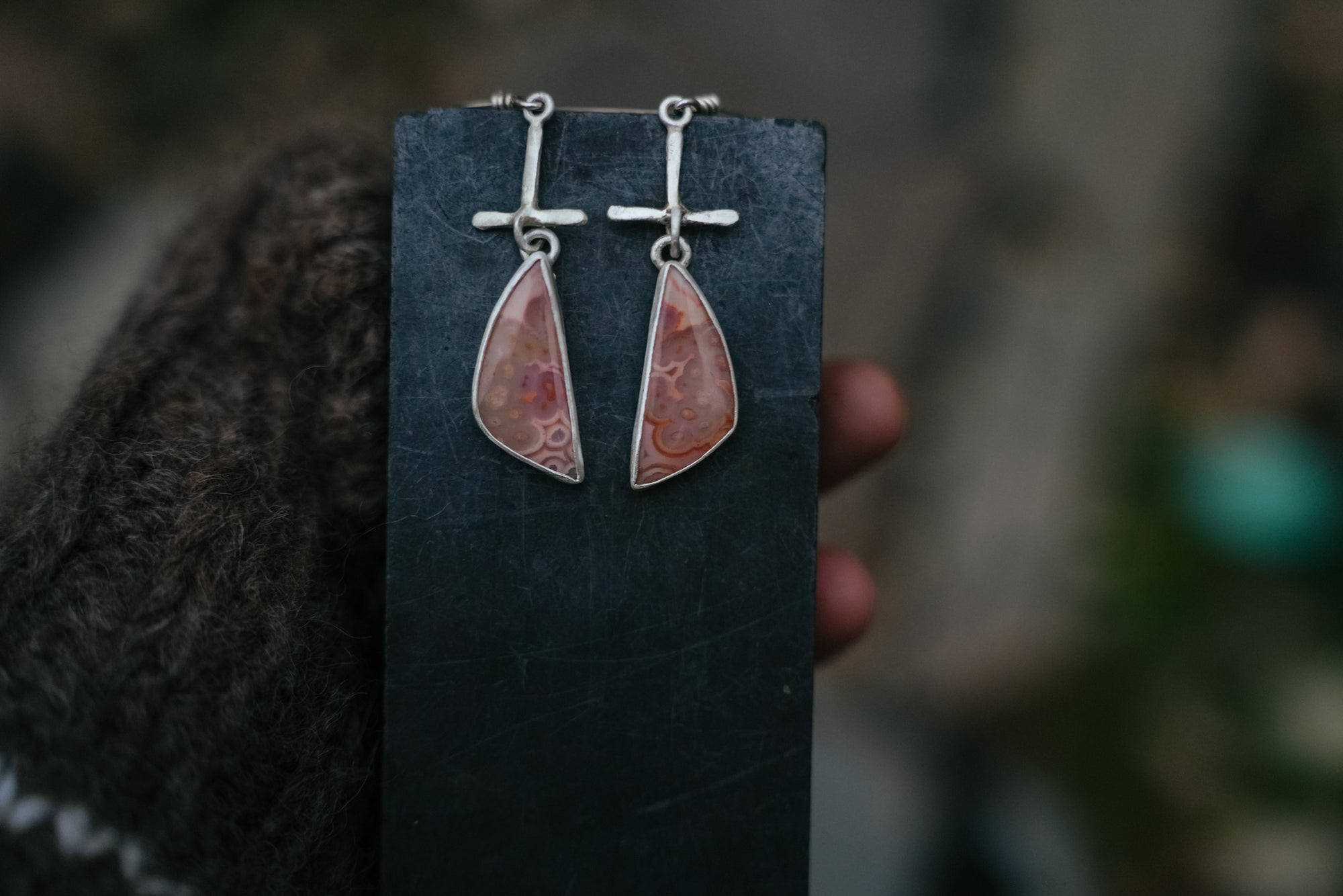 A pair of triangular sterling silver earrings with pink tube agate gemstones dangling from a silver post. They are hanging from a dark card held by a woman’s hand, against a blurred background.