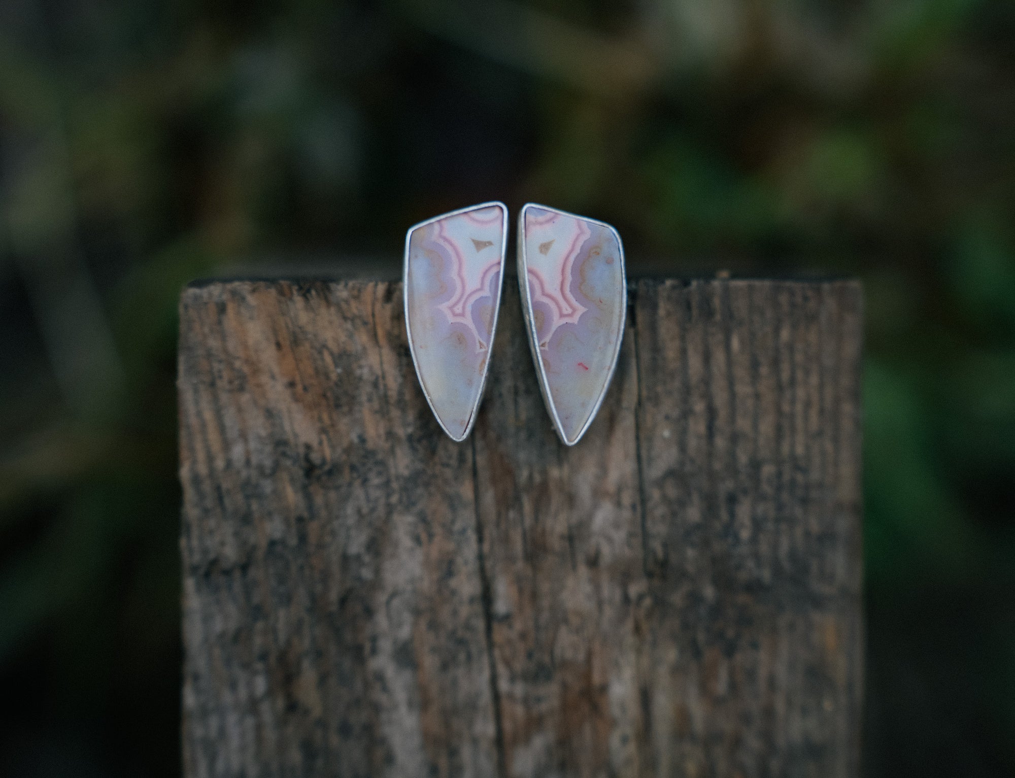 Pair of sterling silver earrings with a pink tube agate gemstones set on a wooden post against a blurred natural background