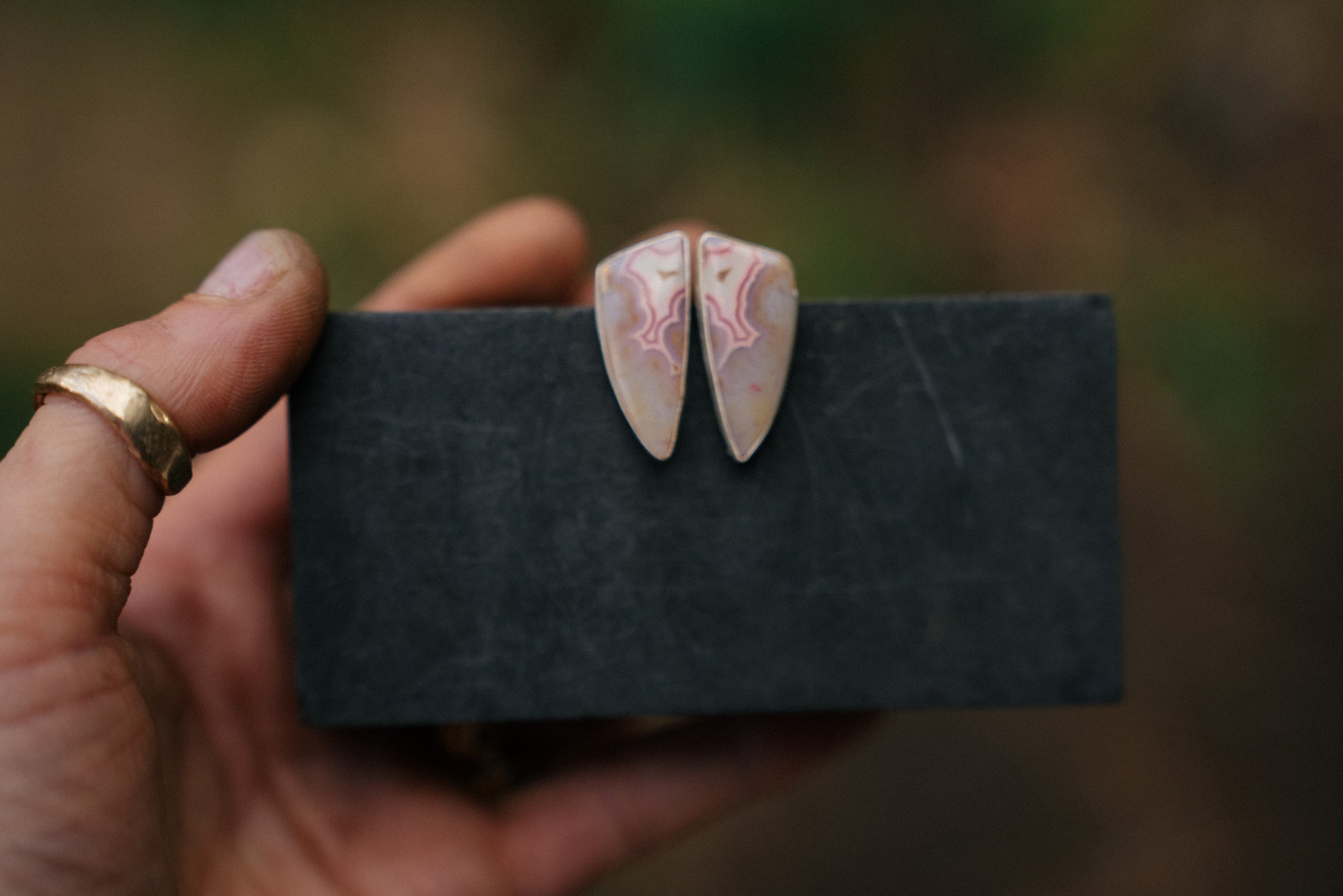 A pair of long triangular-shaped sterling silver post earrings with pink tube agate gemstones. They are hanging from a dark card held by a woman’s hand, against a blurred background.