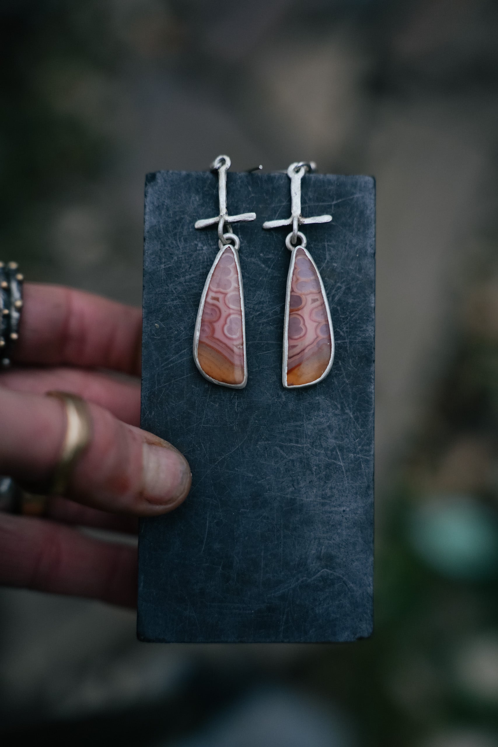 A pair of long triangular-shaped sterling silver post earrings with pink tube agate gemstones. They are hanging from a dark card held by a woman’s hand, against a blurred background.