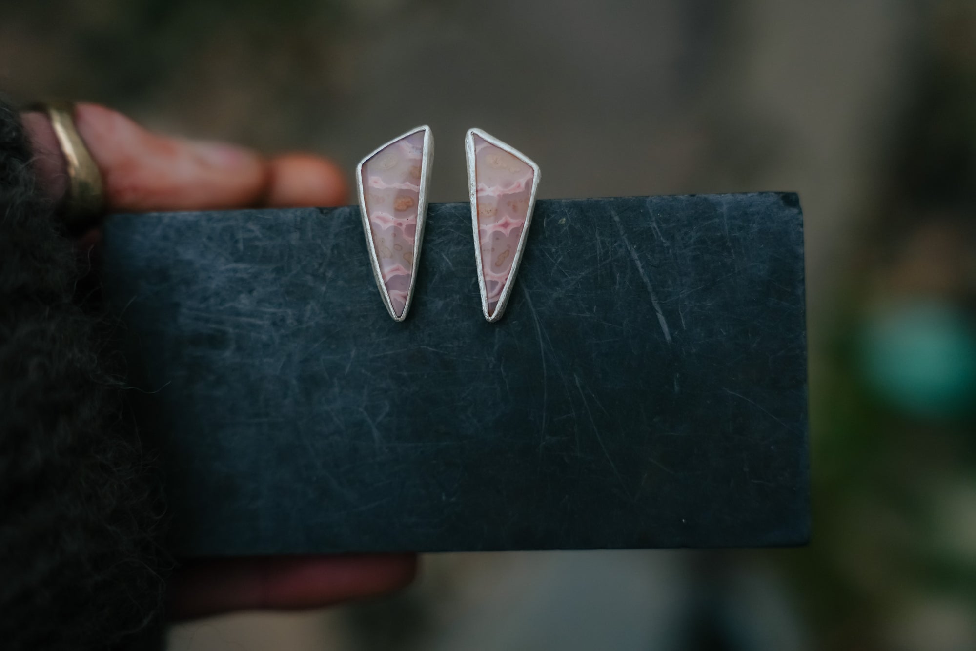 A pair of triangular-shaped sterling silver post earrings with pink tube agate gemstones. They are hanging from a dark card held by a woman’s hand, against a blurred background.
