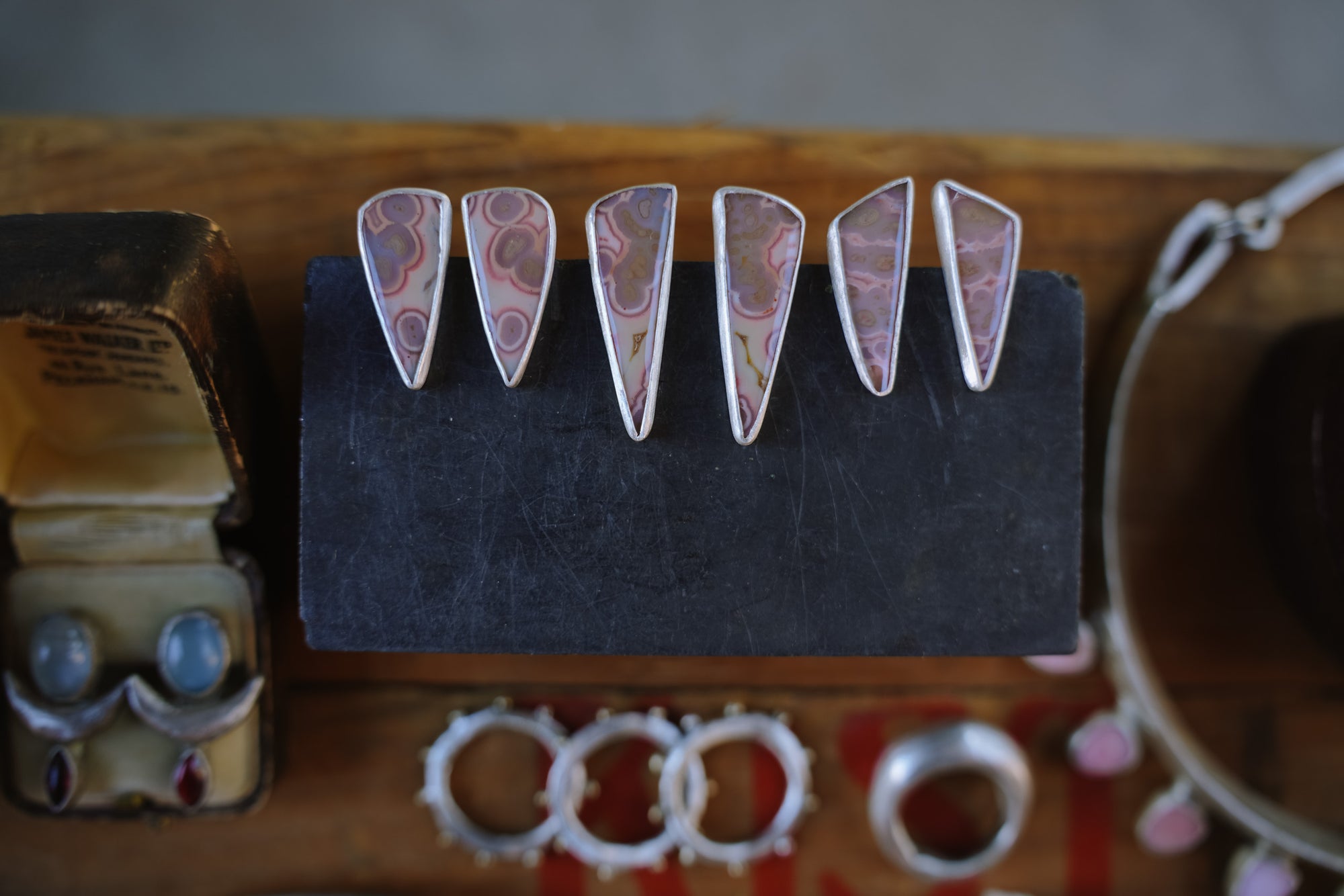 Three pairs of triangular sterling silver earrings with pink tube agate gemstones set on a dark surface on a wooden table with blurred rings and earrings in the background.