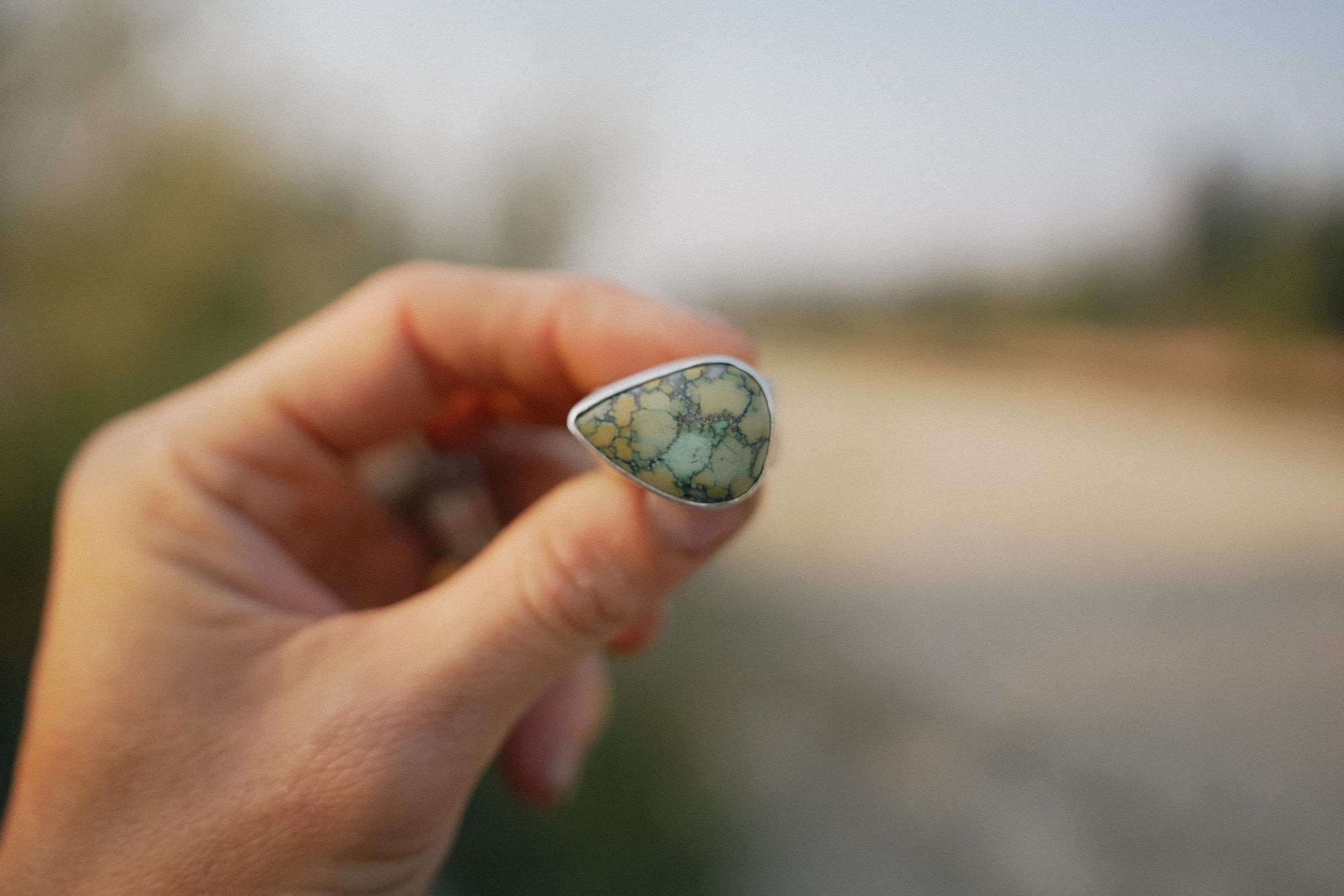 A woman's hand holding a sterling silver and green turquoise ring against a blurred natural background