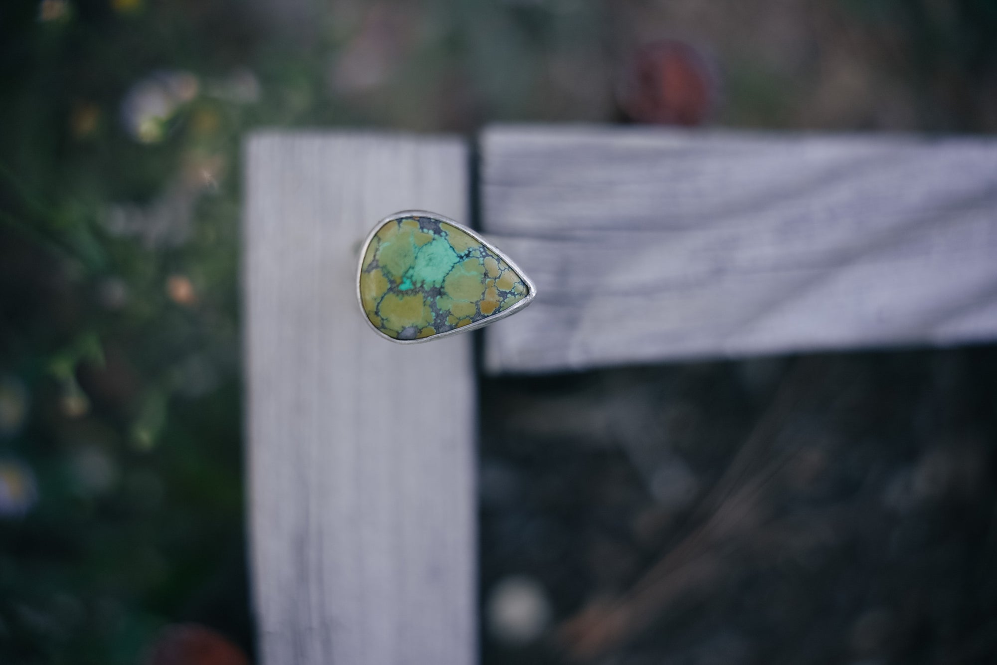 Teardrop-shaped green turquoise and sterling silver ring on a wooden surface against a blurred natural background