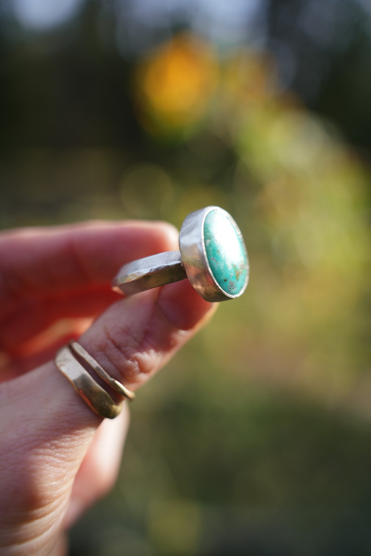 Pool ring, a sterling silver band with an oval turquoise stone in a smooth silver bezel, held up between fingers in side view, with two thin gold bands stacked on the finger below and a blurred green and golden outdoor background.