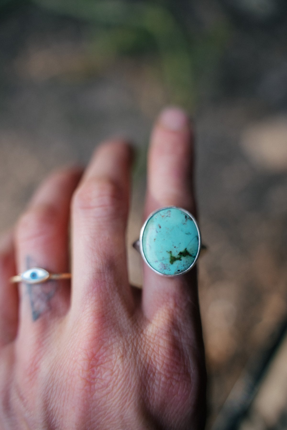 Pool ring, a sterling silver ring with a large oval turquoise stone in a smooth bezel, worn on a raised finger. The pale blue-green stone has dark brown and green matrix veining. A thin gold ring with a small shell eye detail is on the neighboring finger, against a softly blurred earthy background.
