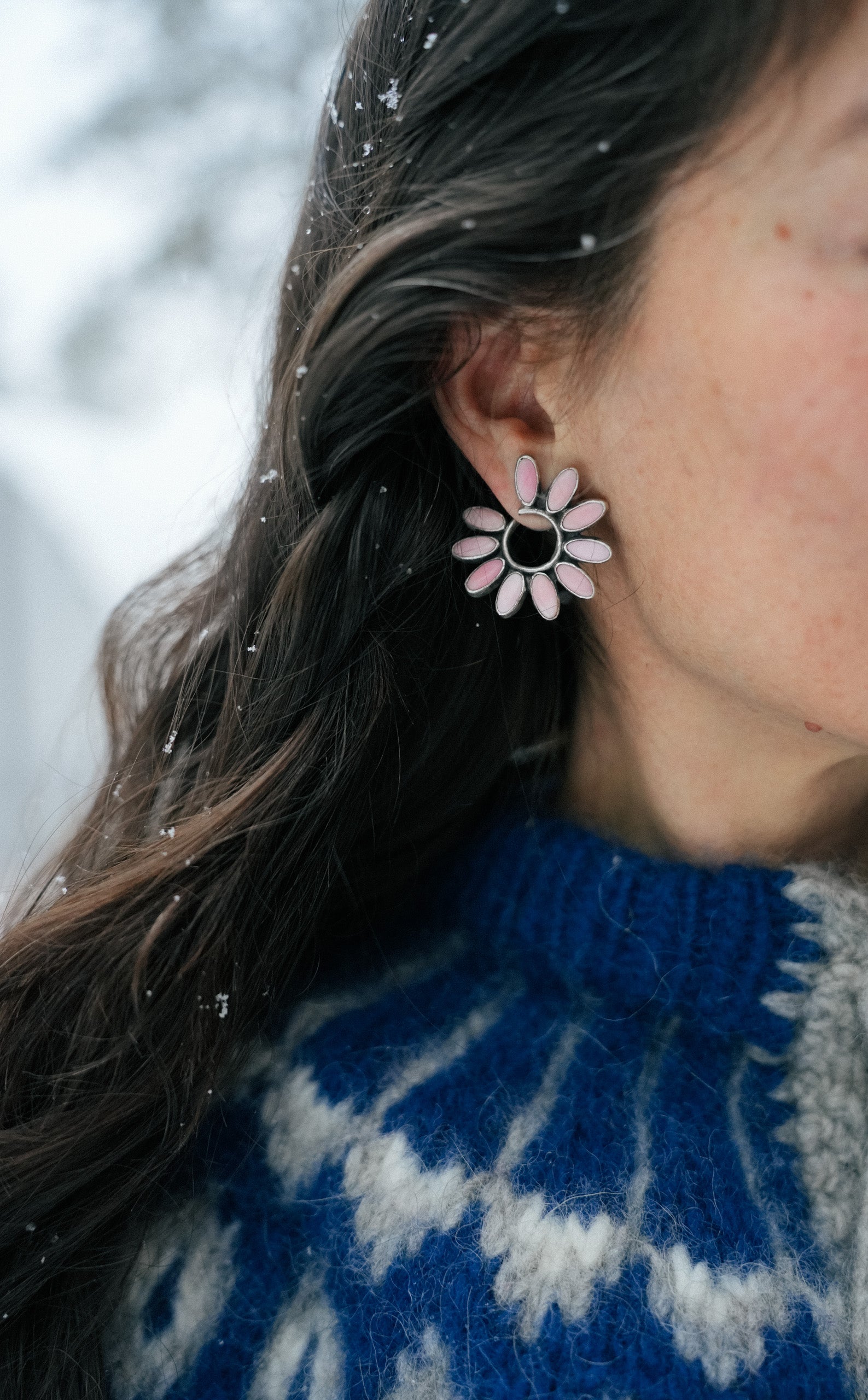 Close-up of a woman with long, dark hair wearing a flower-shaped sterling silver and pink opal earring against a snowy background