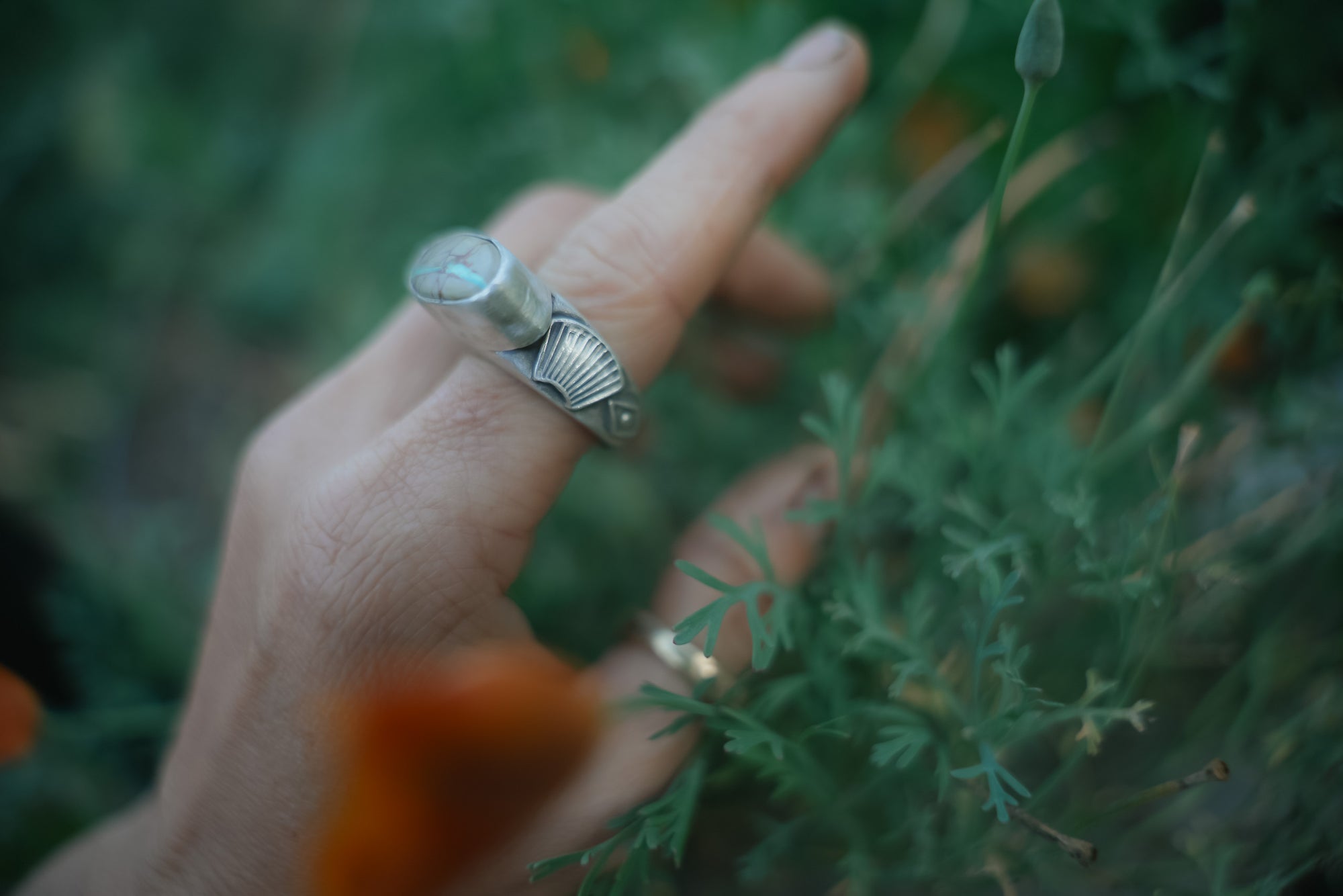 Hand wearing a silver ring with a textured band and a mounted turquoise stone, touching green foliage and orange flowers.