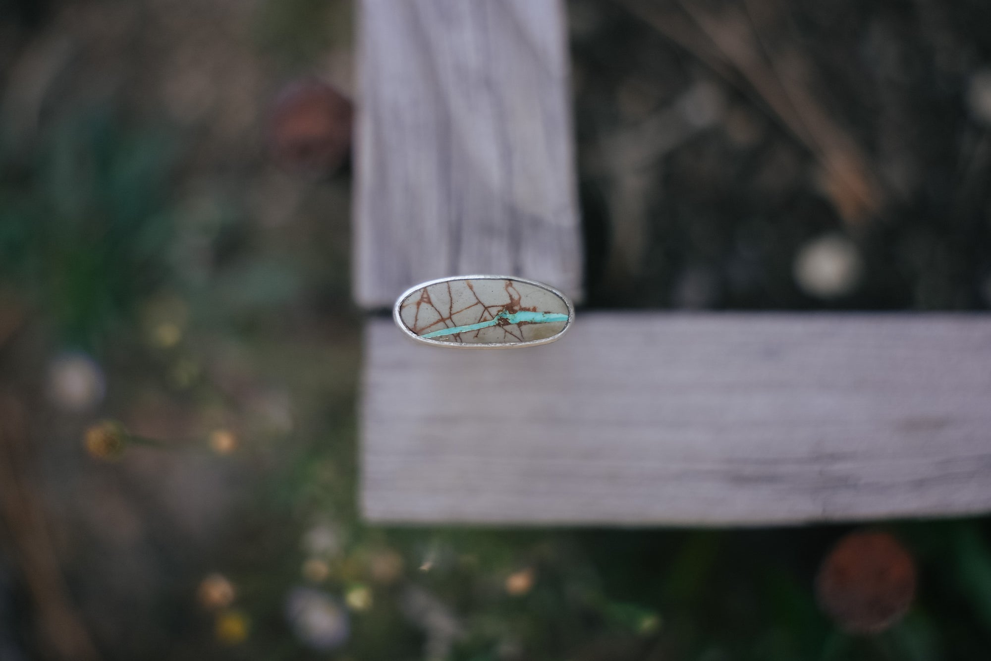 Top view of an oval turquoise stone with thin brown and turquoise lines running through it against a blurred natural background