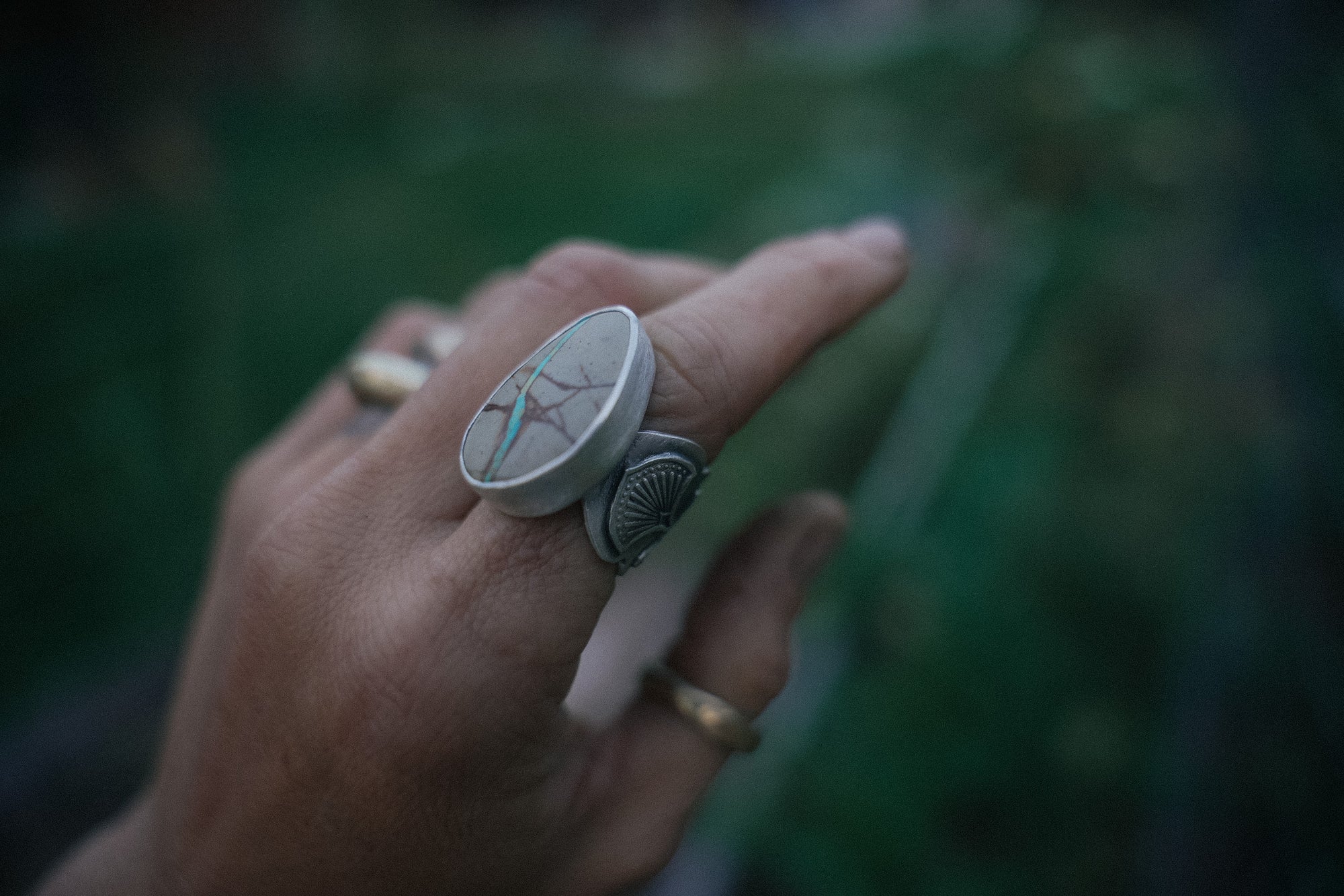 A woman's hand wearing a sterling silver and turquoise ring with a wide, hand-stamped band against a blurred green background