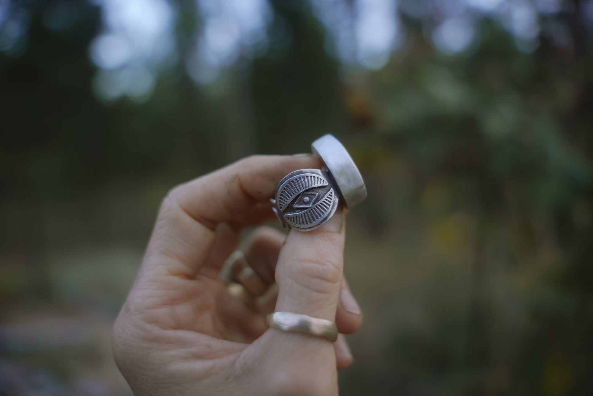 A woman's hand holding a sterling silver ring with a hand-stamped design on the band against a blurred natural background