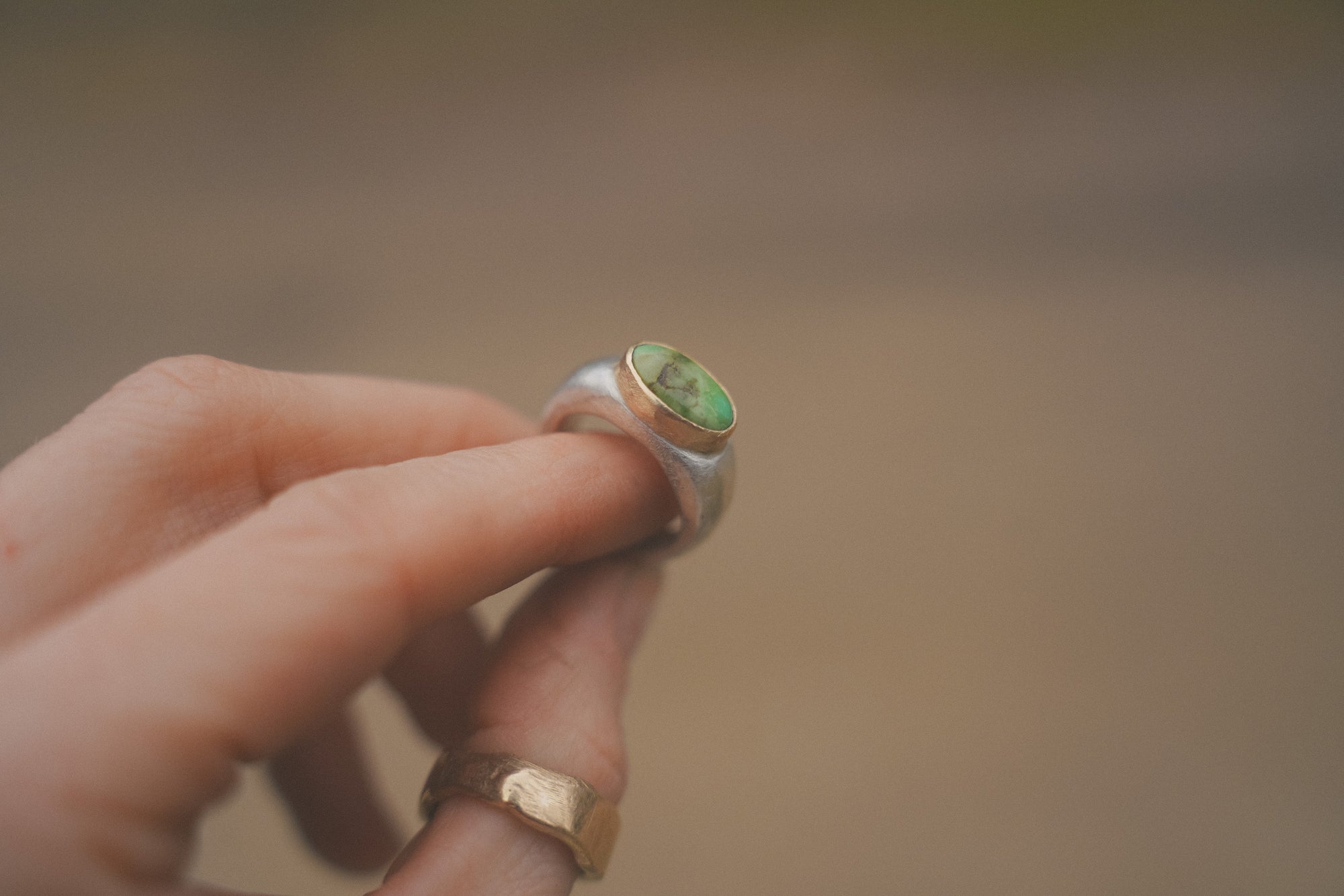 Close-up of a woman's hand holding a sterling silver and 18K gold signet ring with a green turquoise gemstone against a blurred background. She is also wearing a textured gold band on her thumb.