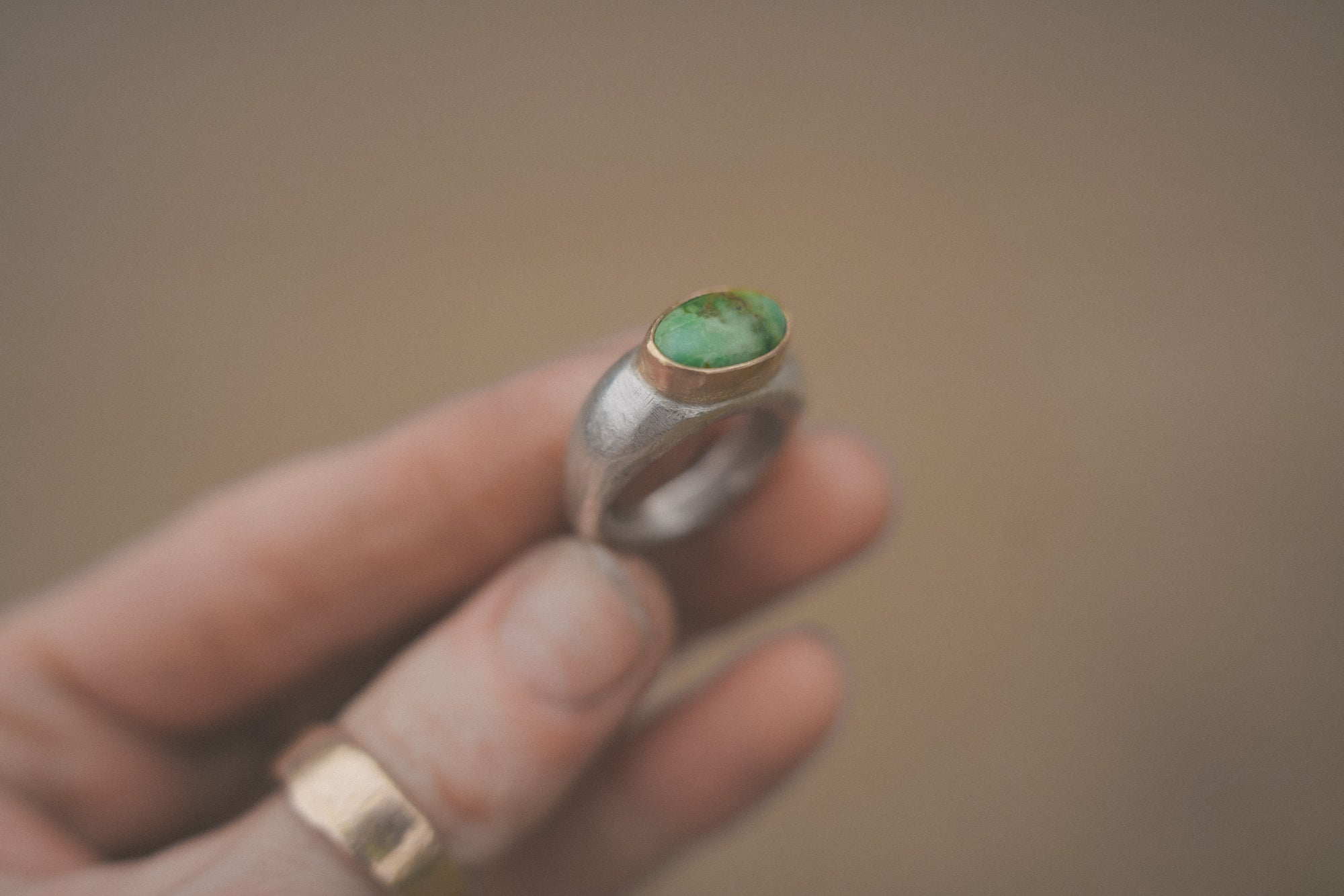 A woman's hand holding a sterling silver and 18K gold signet ring with a green turquoise gemstone against a blurred background.