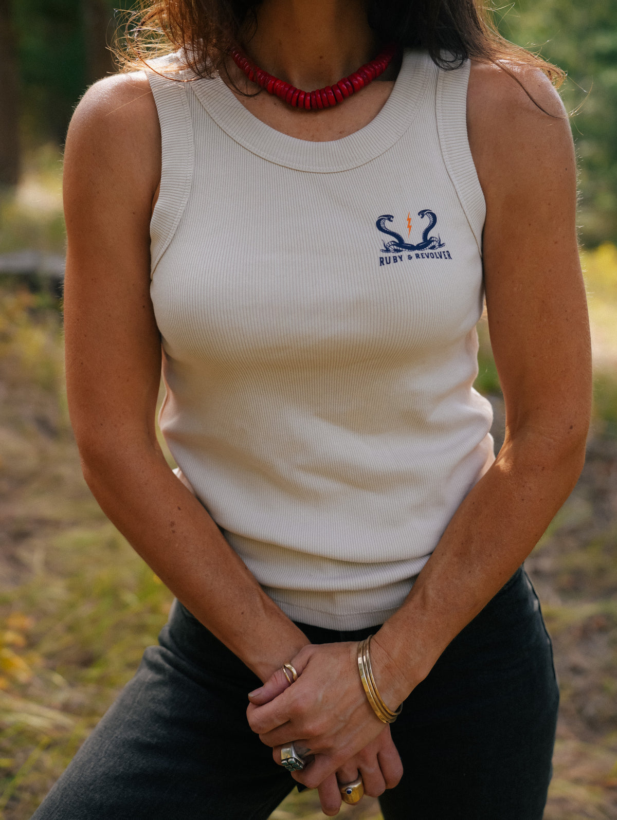 A woman wearing a ribbed off-white tank top featuring a small navy blue graphic on the left chest showing two snakes and a lightning bolt, with the text 'Ruby & Revolver '. She is wearing black pants, photographed outdoors with a blurred natural background.