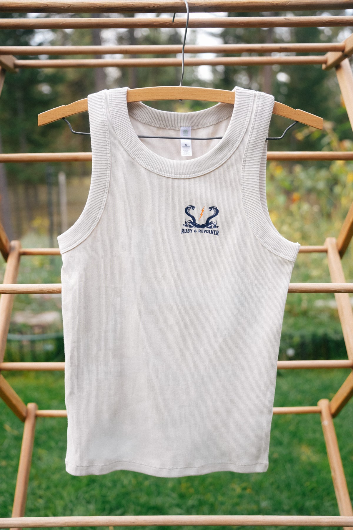 A sleeveless off-white tank top hangs on a wooden hanger on an outdoor wooden rack, featuring a small navy blue graphic on the left chest showing two snakes and a lightning bolt, with the text 'Ruby & Revolver'.