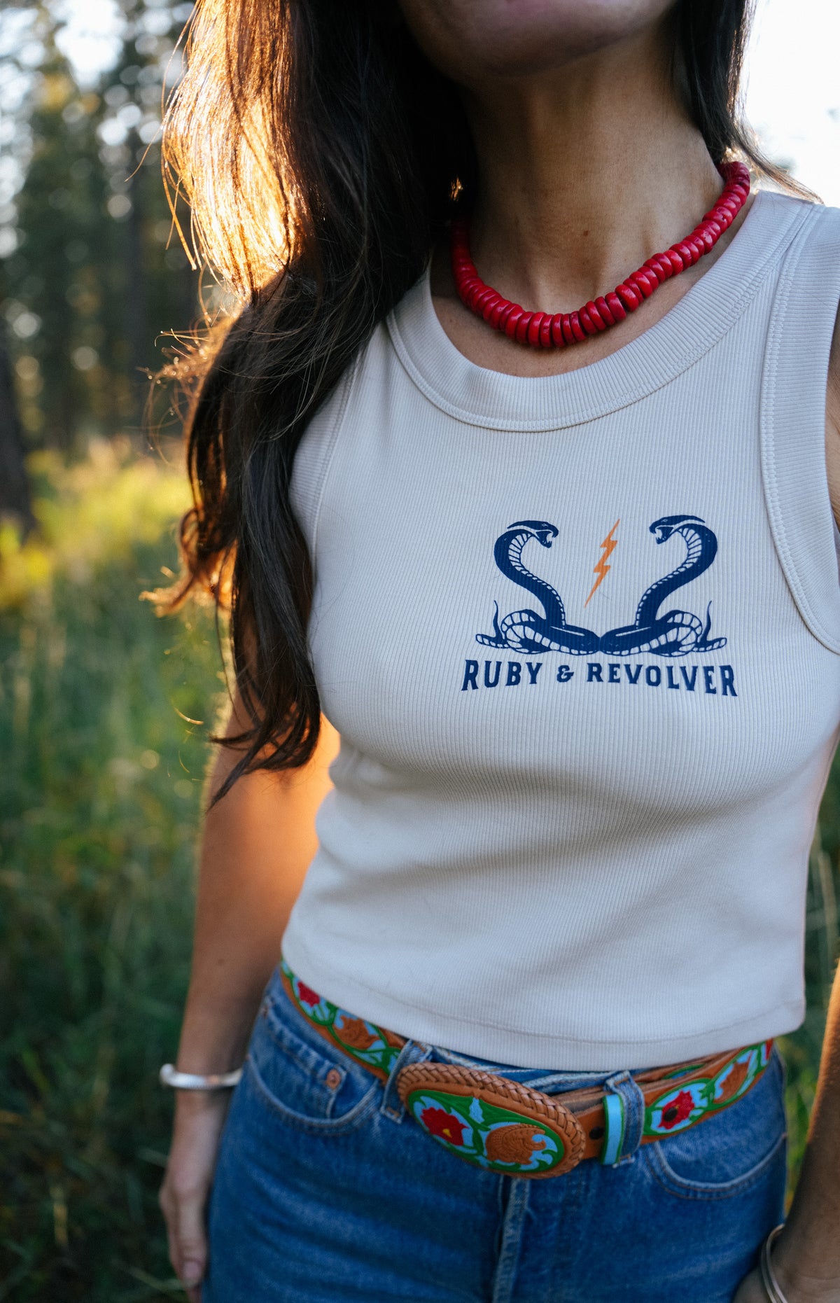 A woman wearing a white ribbed tank top with a blue 'Ruby & Revolver' snake graphic and lightning bolt logo, paired with blue jeans and a colorful belt, in an outdoor setting.