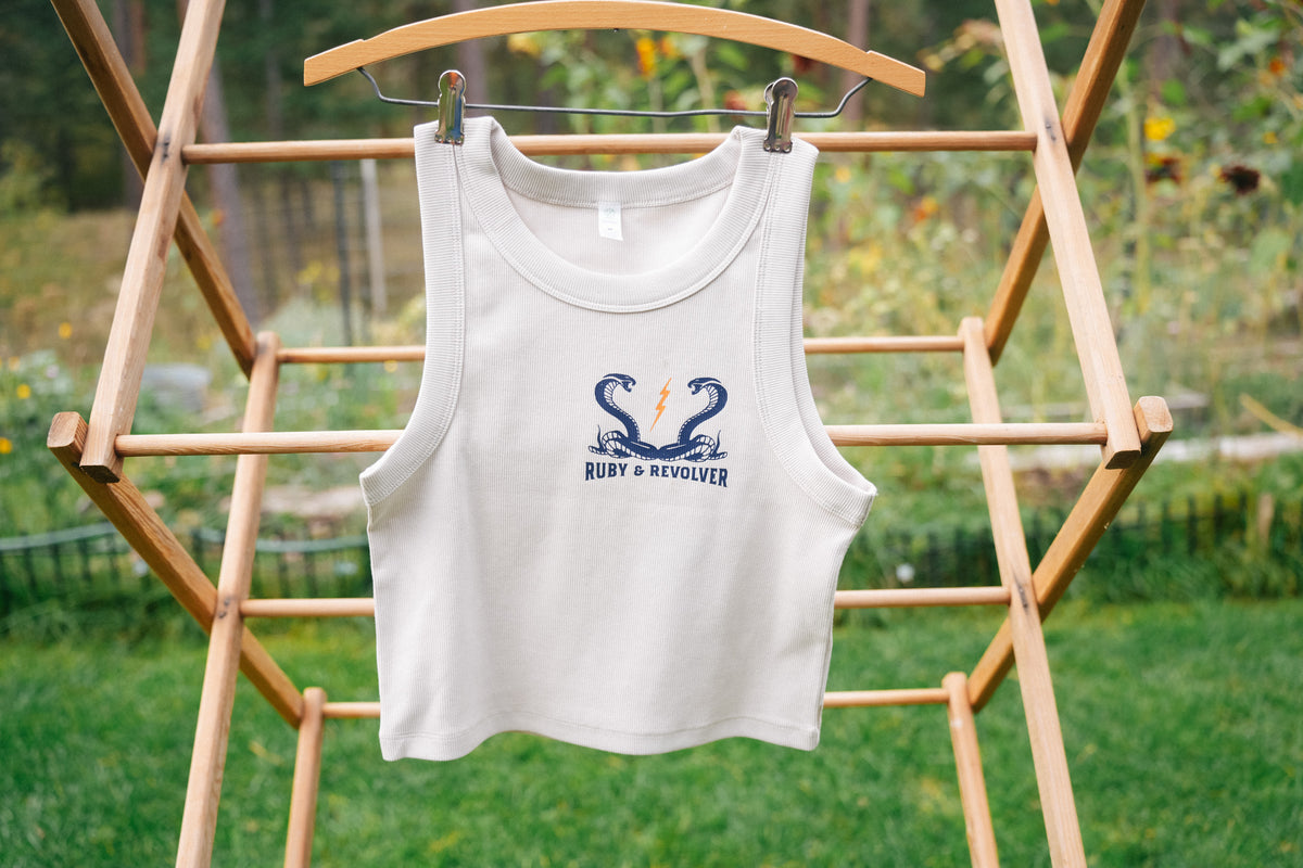 A sleeveless off-white tank top hangs on a wooden hanger on an outdoor wooden rack, featuring a small navy blue graphic on the left chest showing two snakes and a lightning bolt, with the text 'Ruby & Revolver’ outdoors on a grassy lawn.