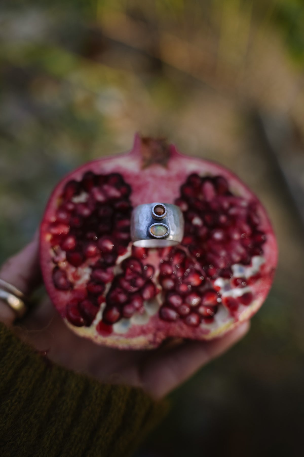 Small Galaxies ring, a wide hammered sterling silver band set with two small stones, a round Montana garnet above an oval Australian opal, resting against the red seeds of a halved pomegranate held in a hand, with a blurred green and brown outdoor background.