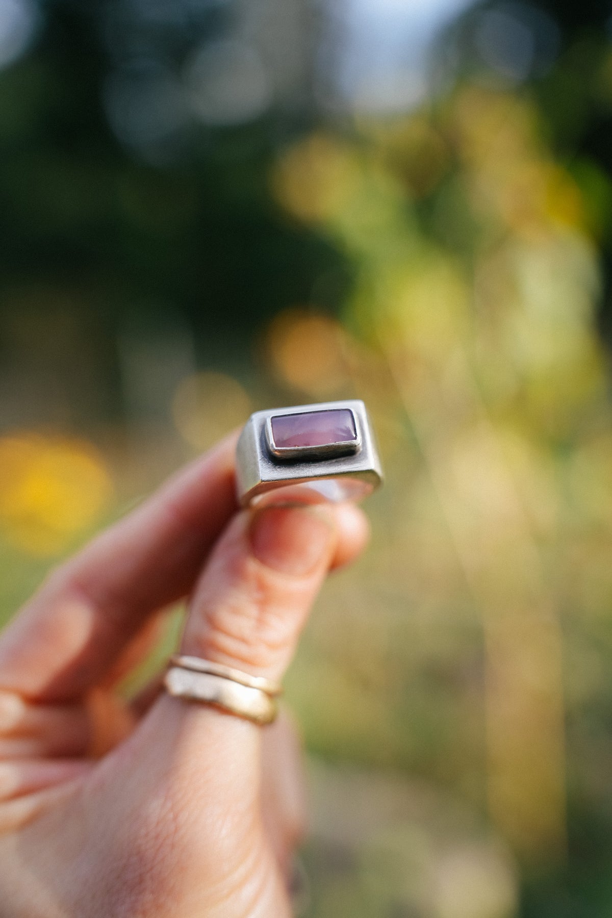 Close-up of a woman's hand holding up a sterling silver signet-style ring with a rectangular, softly rounded Royal Aztec Lace Agate cabochon, set in a smooth silver bezel. This view shows the ring's wide band and its brown-purple gemstone color. Two thin gold bands are stacked below it on her thumb against a blurred green and golden-yellow outdoor background.