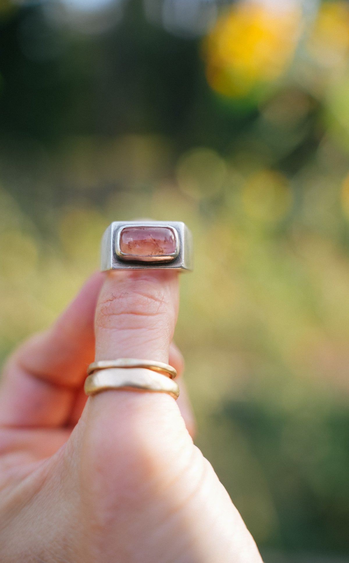 Close-up of a woman's hand holding up a sterling silver signet-style ring with a rectangular, softly rounded Peach Imperial Topaz cabochon, set in a smooth silver bezel. Two thin gold bands are stacked below it on the same finger, against a blurred green and golden-yellow outdoor background.