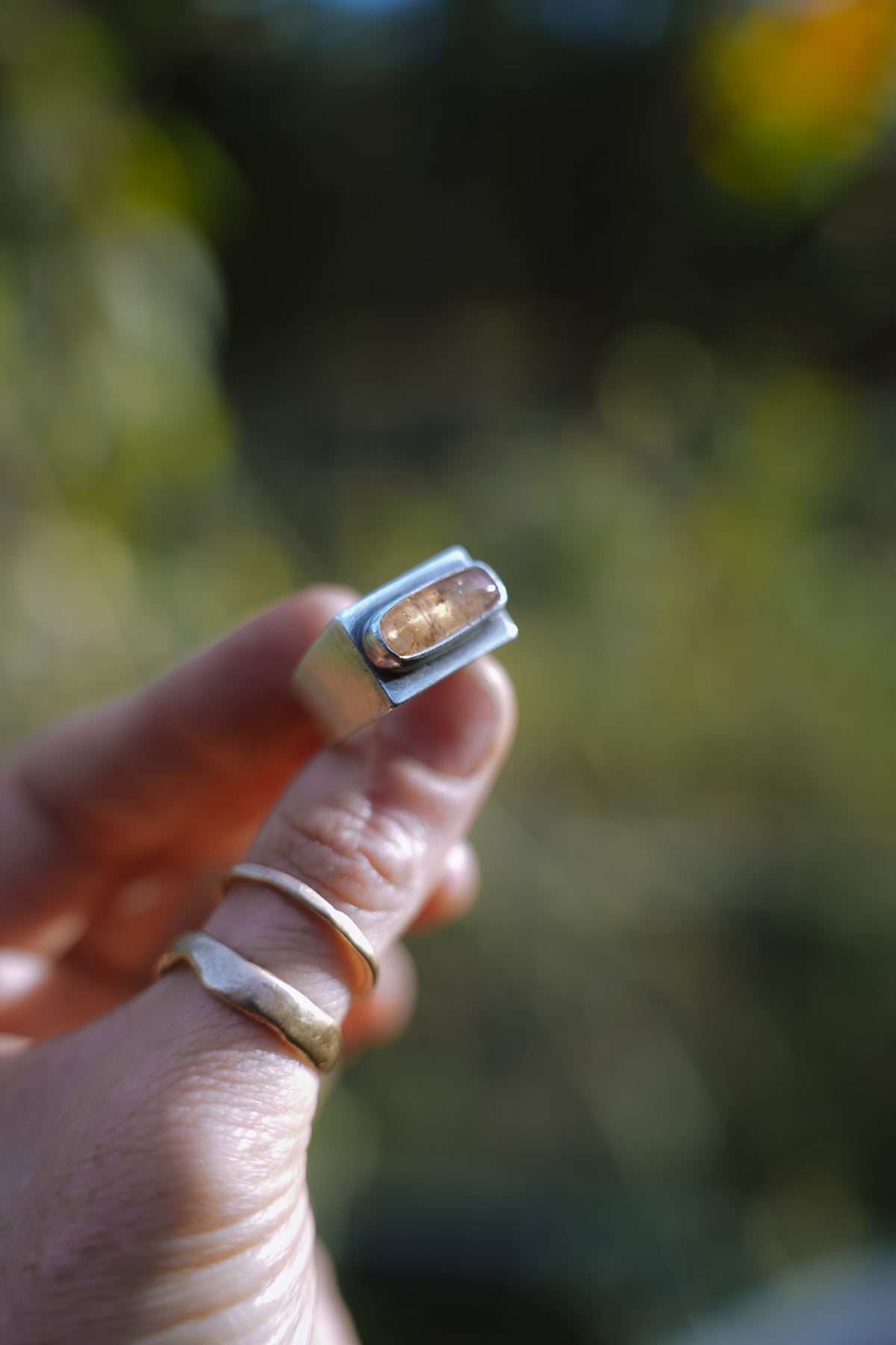 Close-up of a woman's hand holding up a sterling silver signet-style ring with a rectangular, softly rounded Tangerine Imperial Topaz cabochon, set in a smooth silver bezel. This is a side-view of the ring showing the wide band. Two thin gold bands are stacked below it on her thumb against a blurred green and golden-yellow outdoor background.