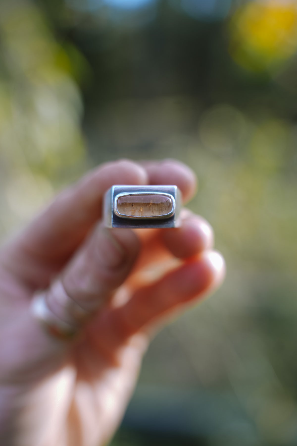 Close-up of a woman's hand holding up a sterling silver signet-style ring with a rectangular, softly rounded Tangerine Imperial Topaz cabochon, set in a smooth silver bezel. This is a top-view of the ring. Two thin gold bands are stacked below it on her thumb against a blurred green and golden-yellow outdoor background.