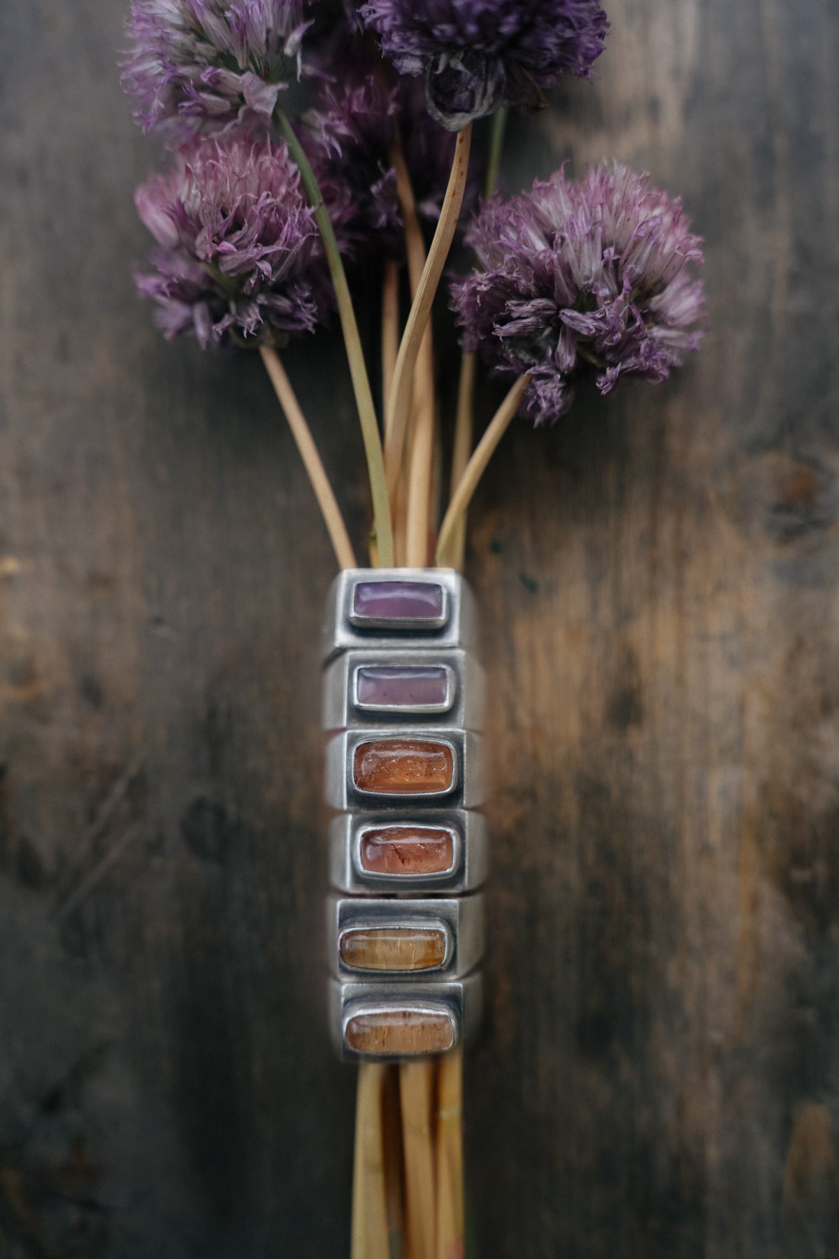 Six sterling silver signet rings with rectangular cabochon stones stacked in a vertical column on a dark, weathered wood surface, with a bundle of dried purple chive blossoms above them. The stones run from lavender and pale purple at the top to peachy-pink, orange, and golden tones at the bottom.