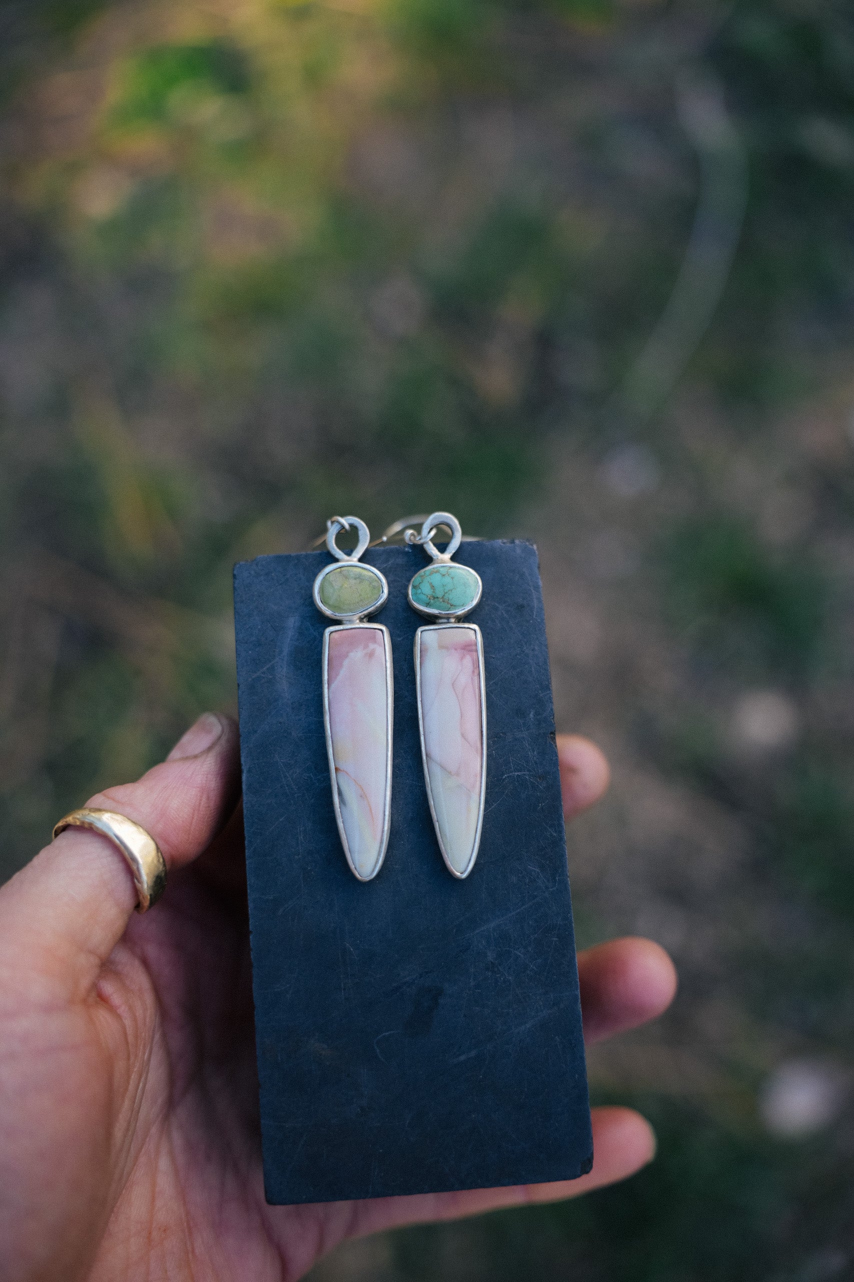 A woman's hand holding a blue velvet card onto which a pair of sterling silver, turquoise, and willow jasper dangler earrings are attached. The natural background is blurred.