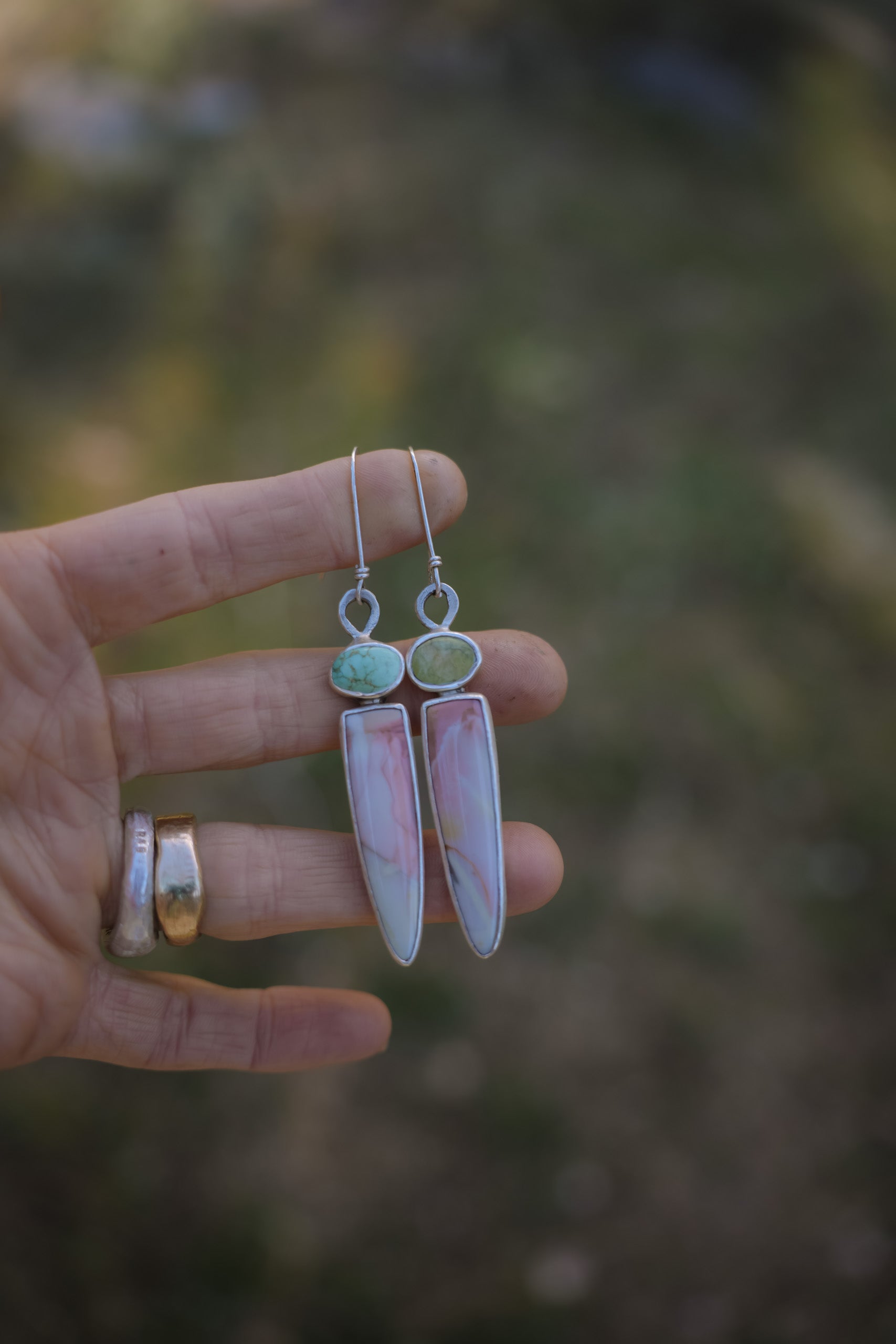 A woman's hand holding a pair of sterling silver, turquoise, and willow jasper dangler earrings earrings. The turquoise gemstones are light green and light blue. The spike-shaped willow jasper gemstone are white with swirls of of pale orange and terracotta colors. The natural background is blurred.