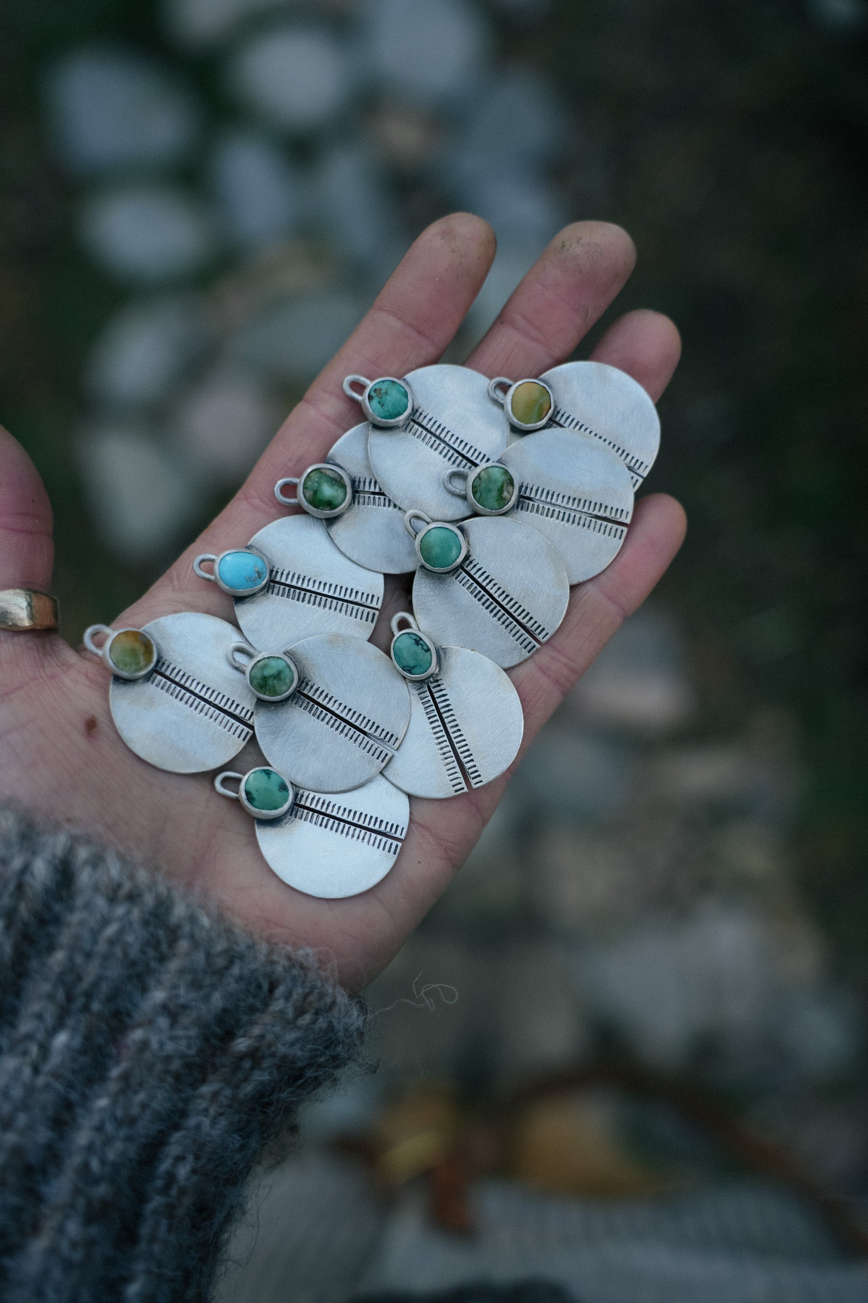 A woman’s hand holding five pairs of hand-stamped sterling silver circular earrings with green and blue turquoise gemstones on a dark background.