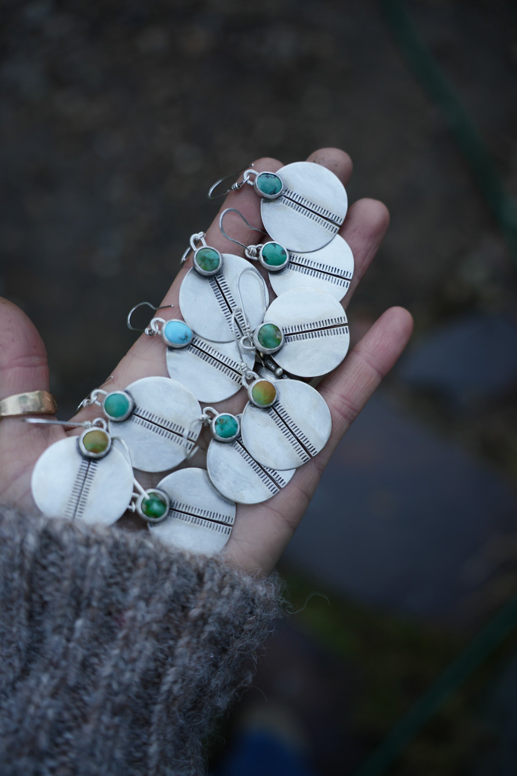 A woman’s hand holding five pairs of hand-stamped sterling silver circular earrings with green and blue turquoise gemstones on a dark background.